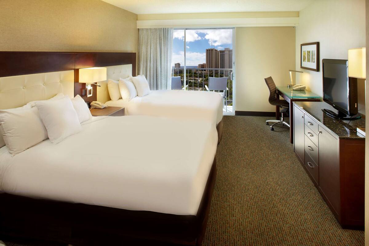 A hotel room with two beds, a desk with a computer, a TV, and a view of the cityscape.