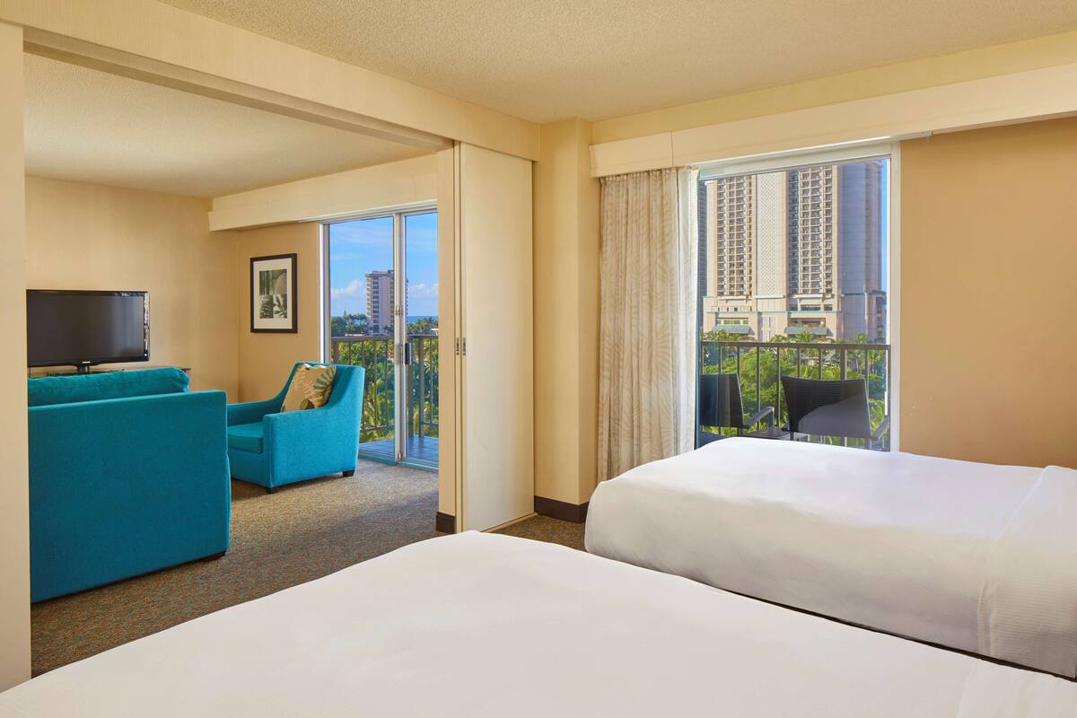 A hotel room with two beds, a sofa, a TV, and a balcony with a view of buildings.