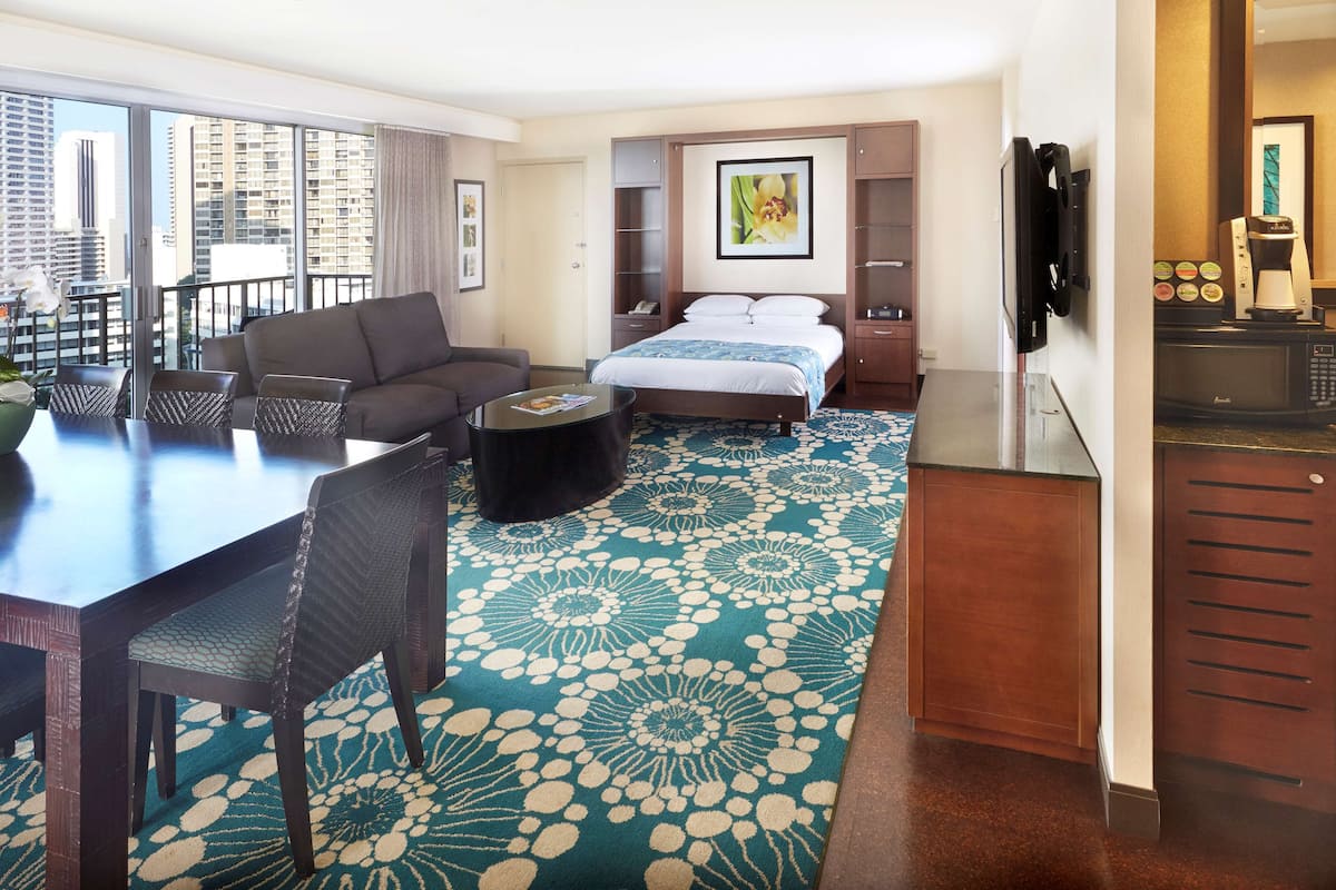 A hotel room with a bed, a sofa, a dining table with chairs, a coffee machine, and a view of the city.