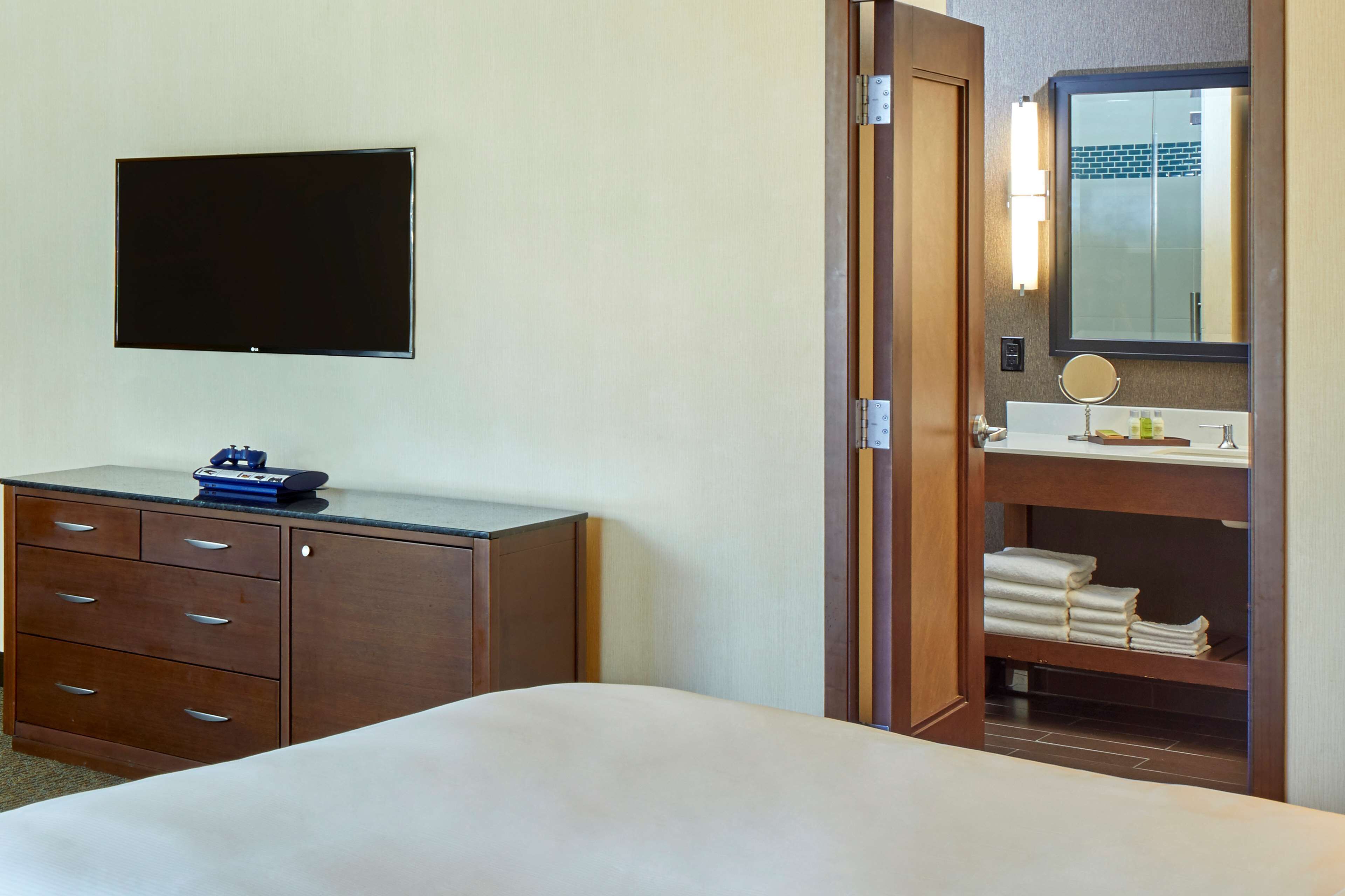 A hotel room with a bed, a wooden dresser with drawers, a flat-screen TV mounted on the wall, and a bathroom with a sink, mirror, and towel rack.