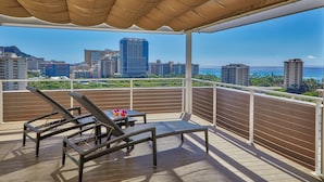 A balcony with two lounge chairs and a table, overlooking a cityscape and the ocean.