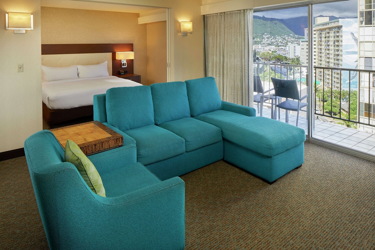 A hotel room with a large bed, a teal sofa, a small wooden table, and a balcony with a city view.