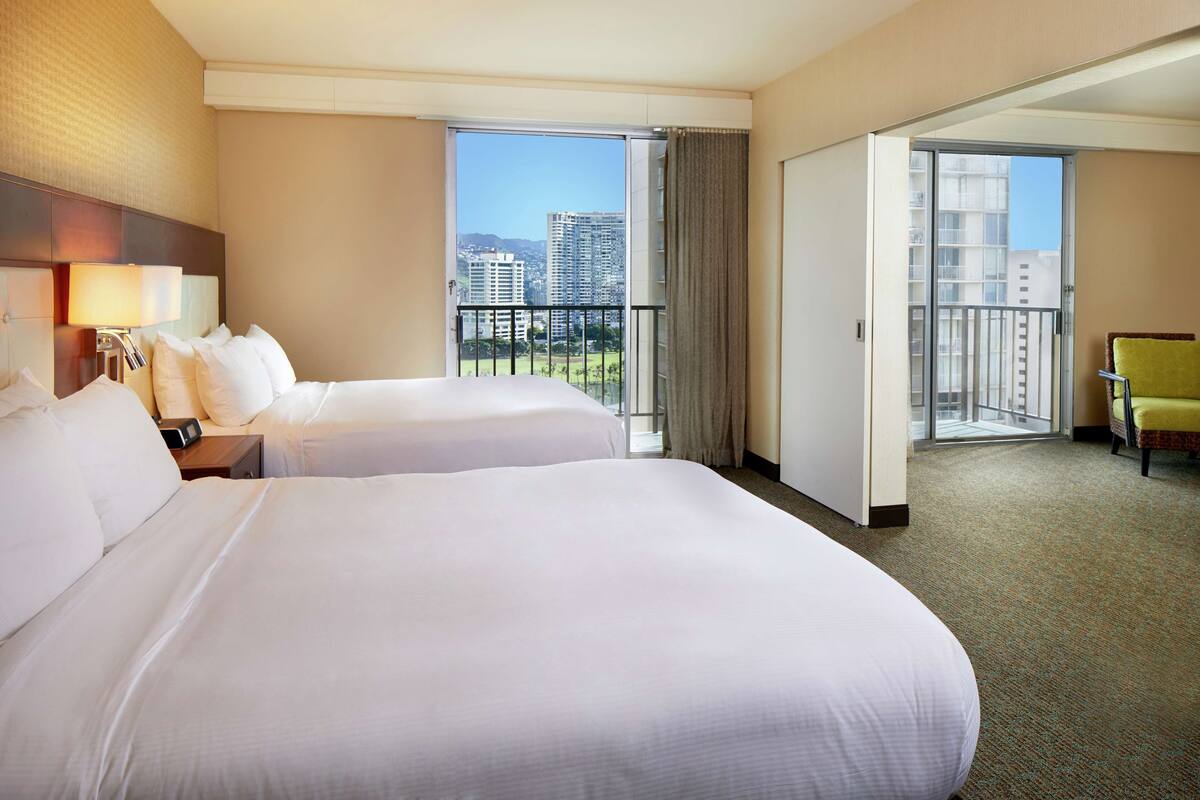 A hotel room with two beds, a nightstand, a lamp, a chair, and a view of the city through large windows.