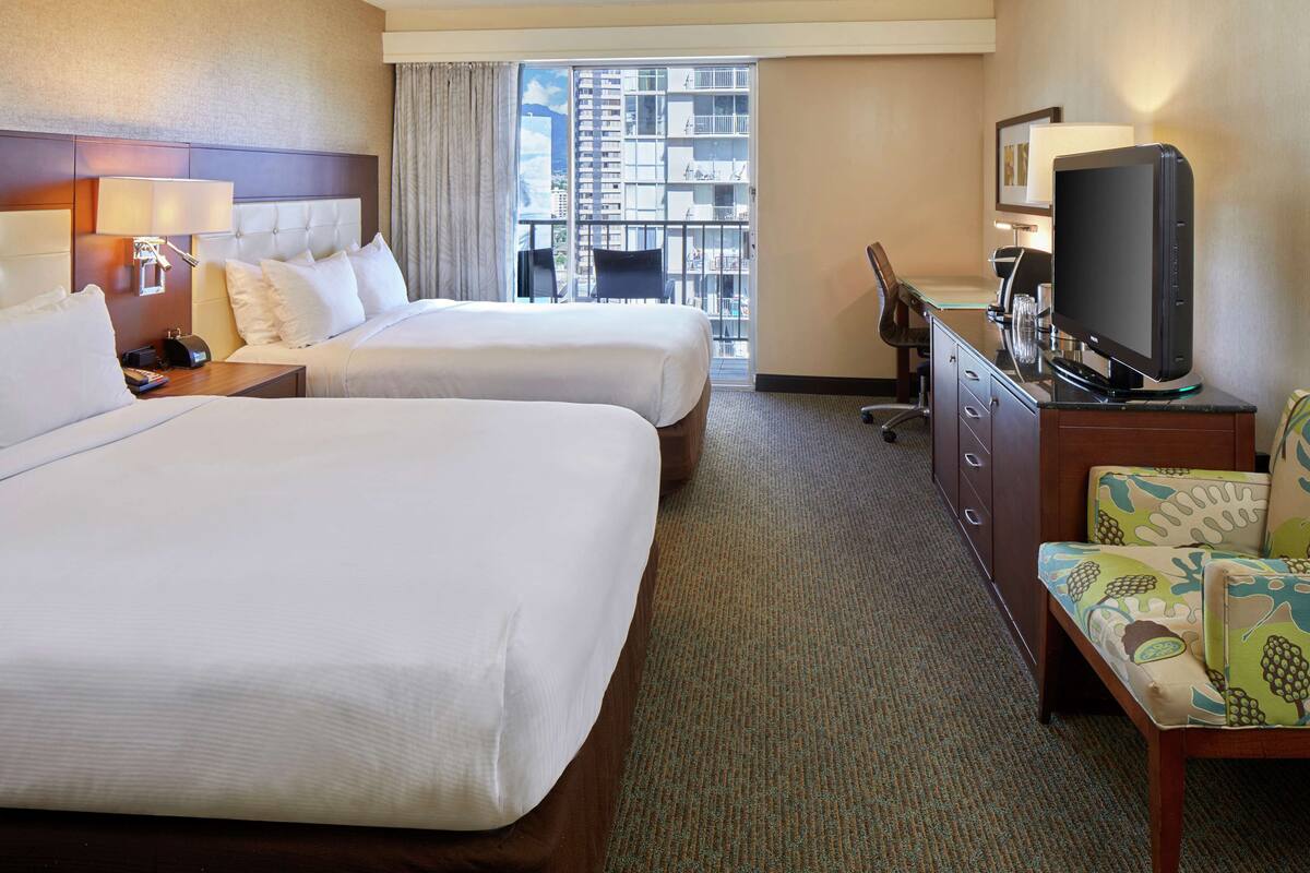 A hotel room with two beds, a television, a desk, and a view of the city through the window.
