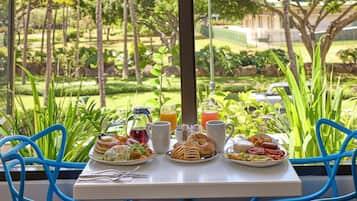 A table set with breakfast items and drinks, overlooking a well-maintained garden with trees and a fence.