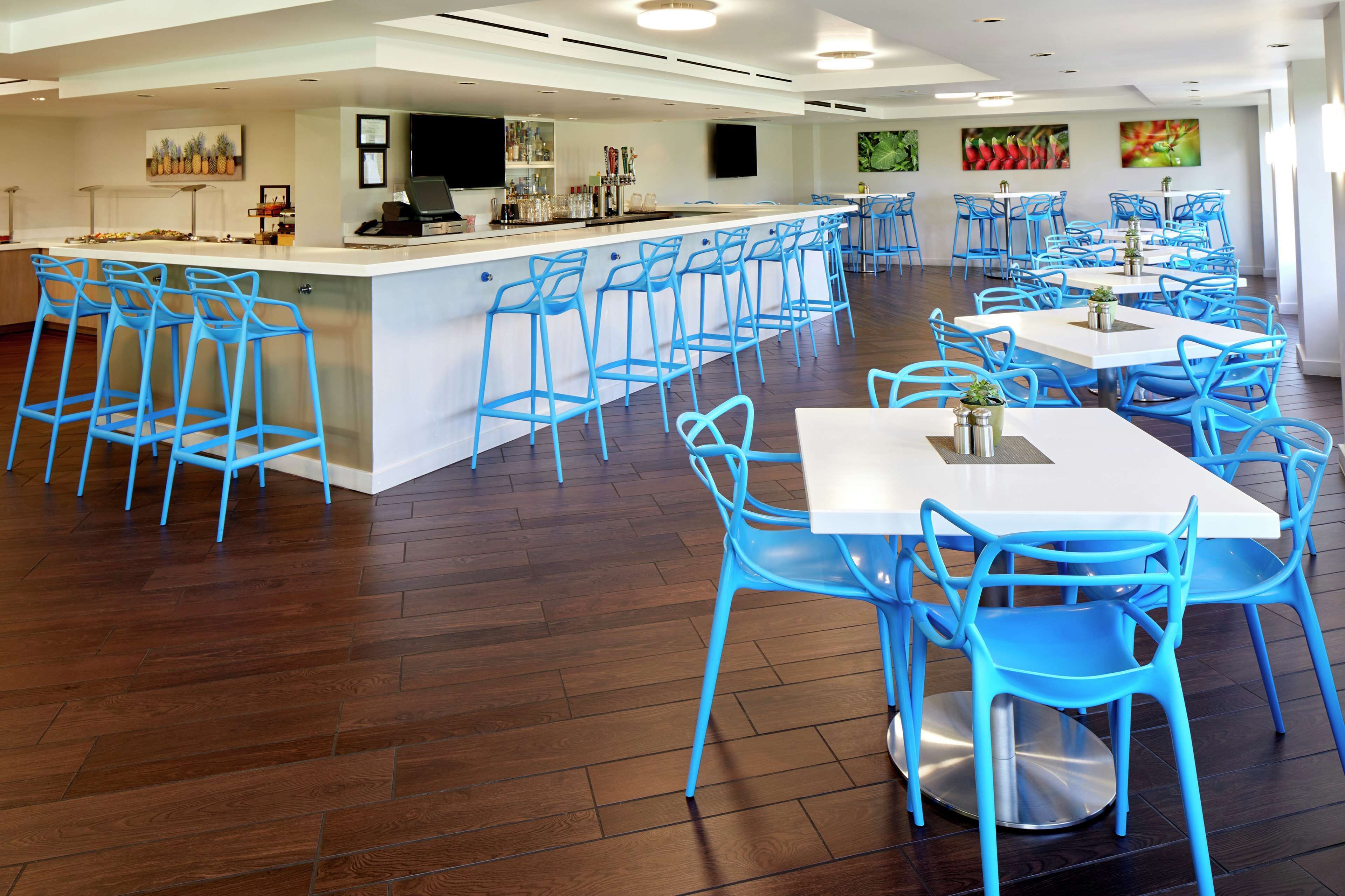 A spacious restaurant with blue chairs and white tables, a bar area, and artwork on the walls.