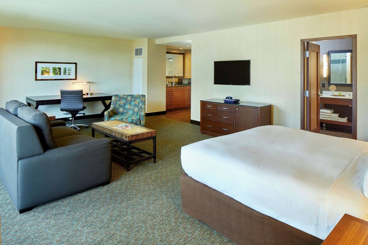 A hotel room with a bed, a desk with a chair, a small table, a sofa, a TV, and a bathroom visible through an open door.