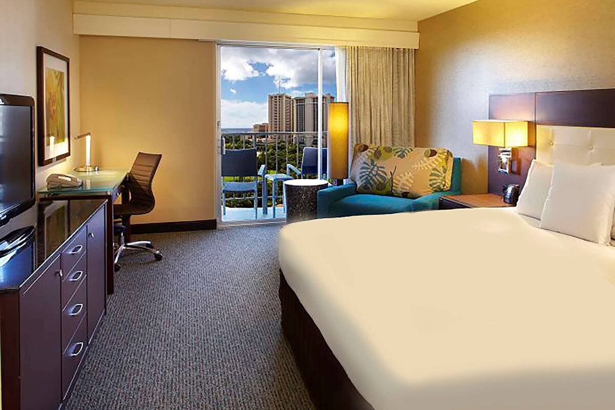A hotel room with a large bed, a desk with a computer, a chair, a sofa, and a balcony with a city view.