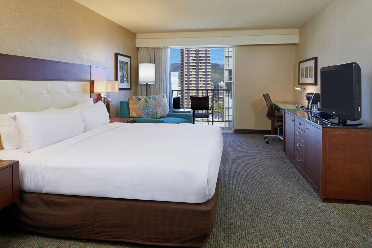 A hotel room with a large bed, a desk with a computer, a chair, a TV, and a view of a building and mountains through the window.