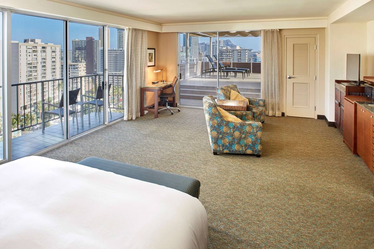 A hotel room with a large bed, a desk with a chair, a sofa, and a view of the city through large windows.
