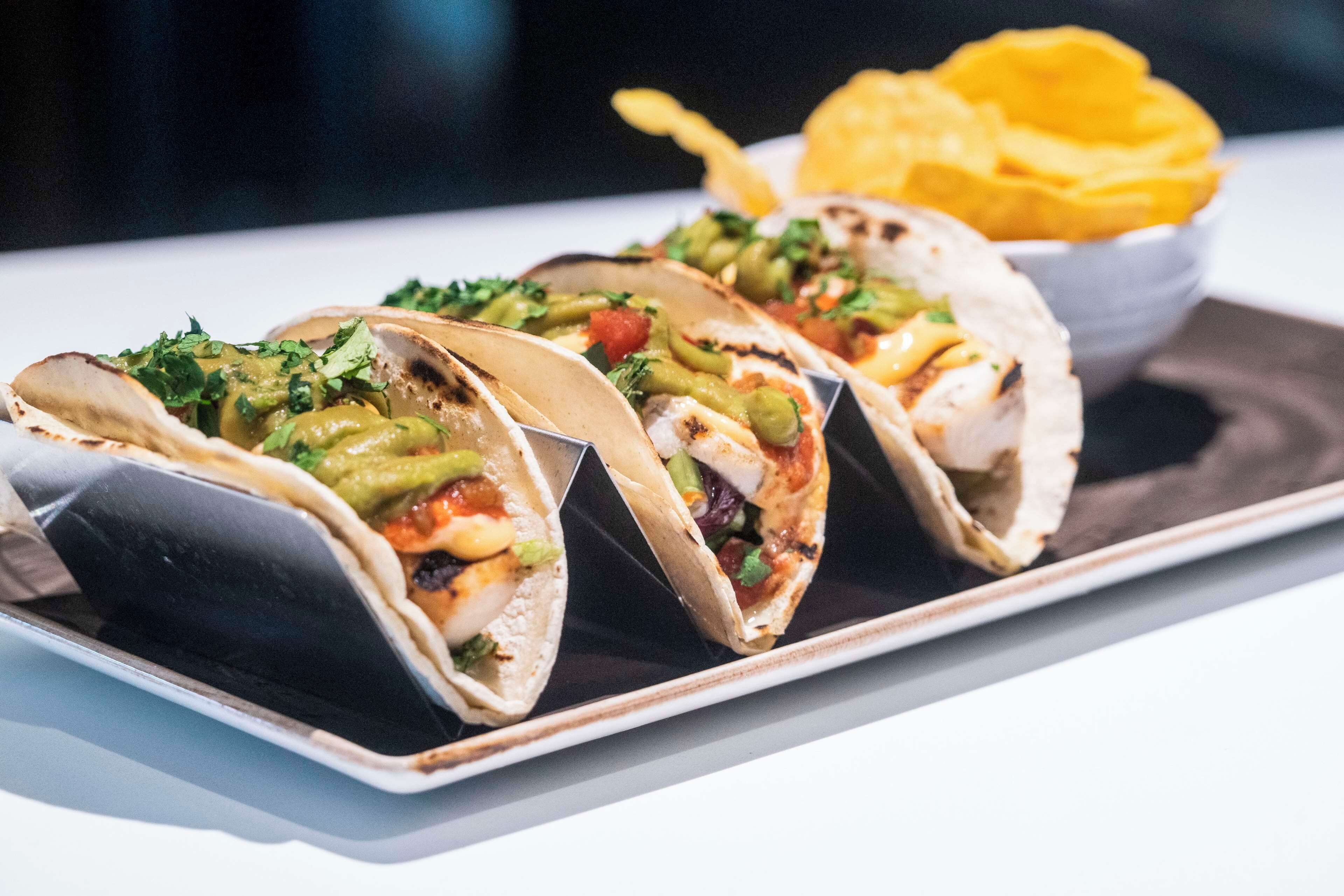 Three tacos with various toppings on a plate, accompanied by a bowl of chips.