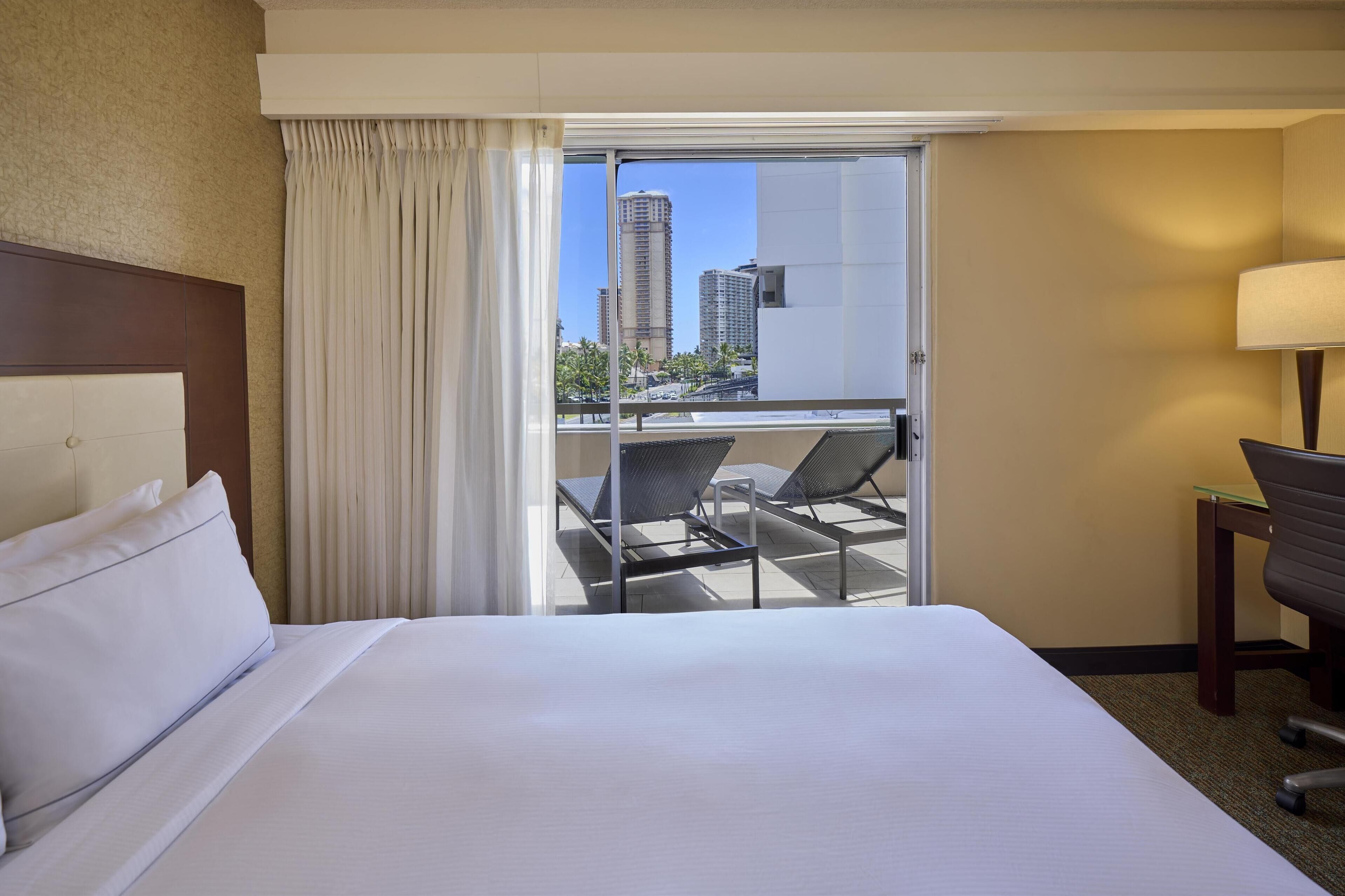 Camera d'albergo con un letto, una scrivania, una sedia e un balcone che si affaccia su alti edifici.