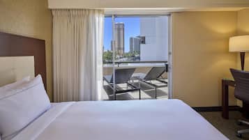 Camera d'albergo con un letto, una scrivania, una sedia e un balcone che si affaccia su alti edifici.