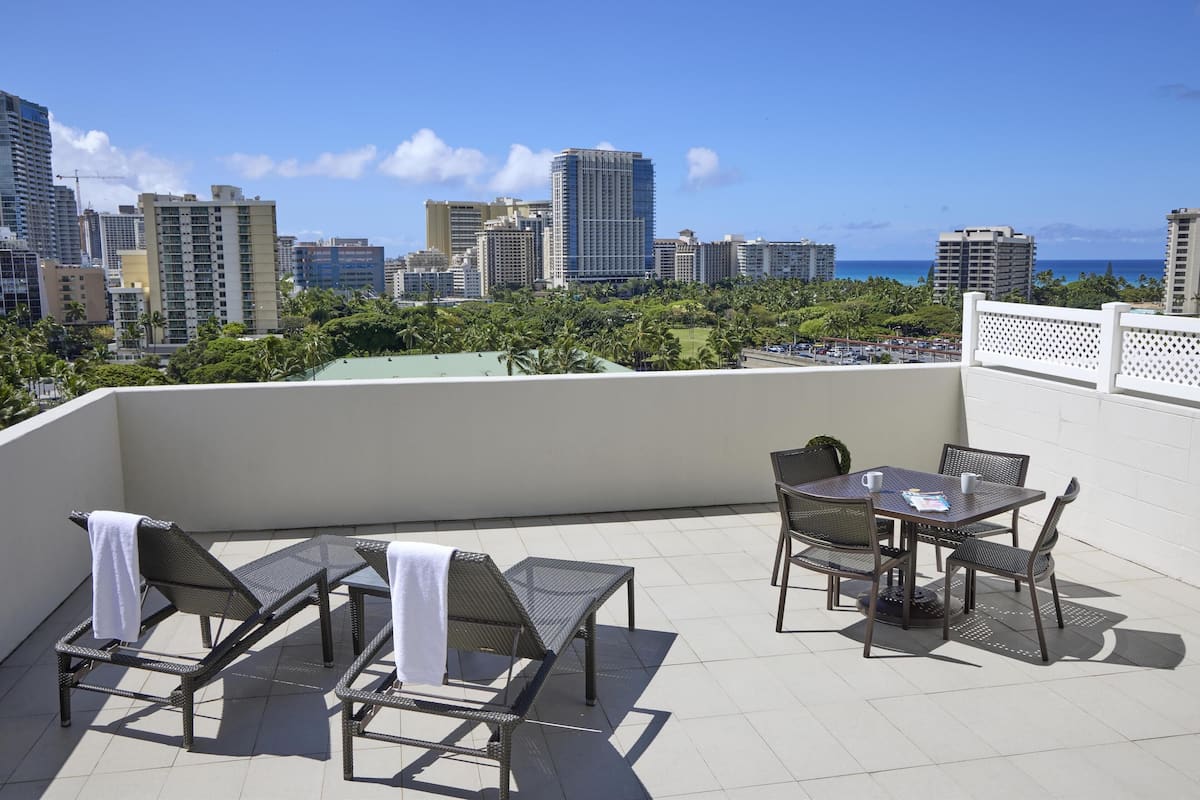 A rooftop terrace with lounge chairs, a table, and chairs, overlooking a cityscape and the ocean.