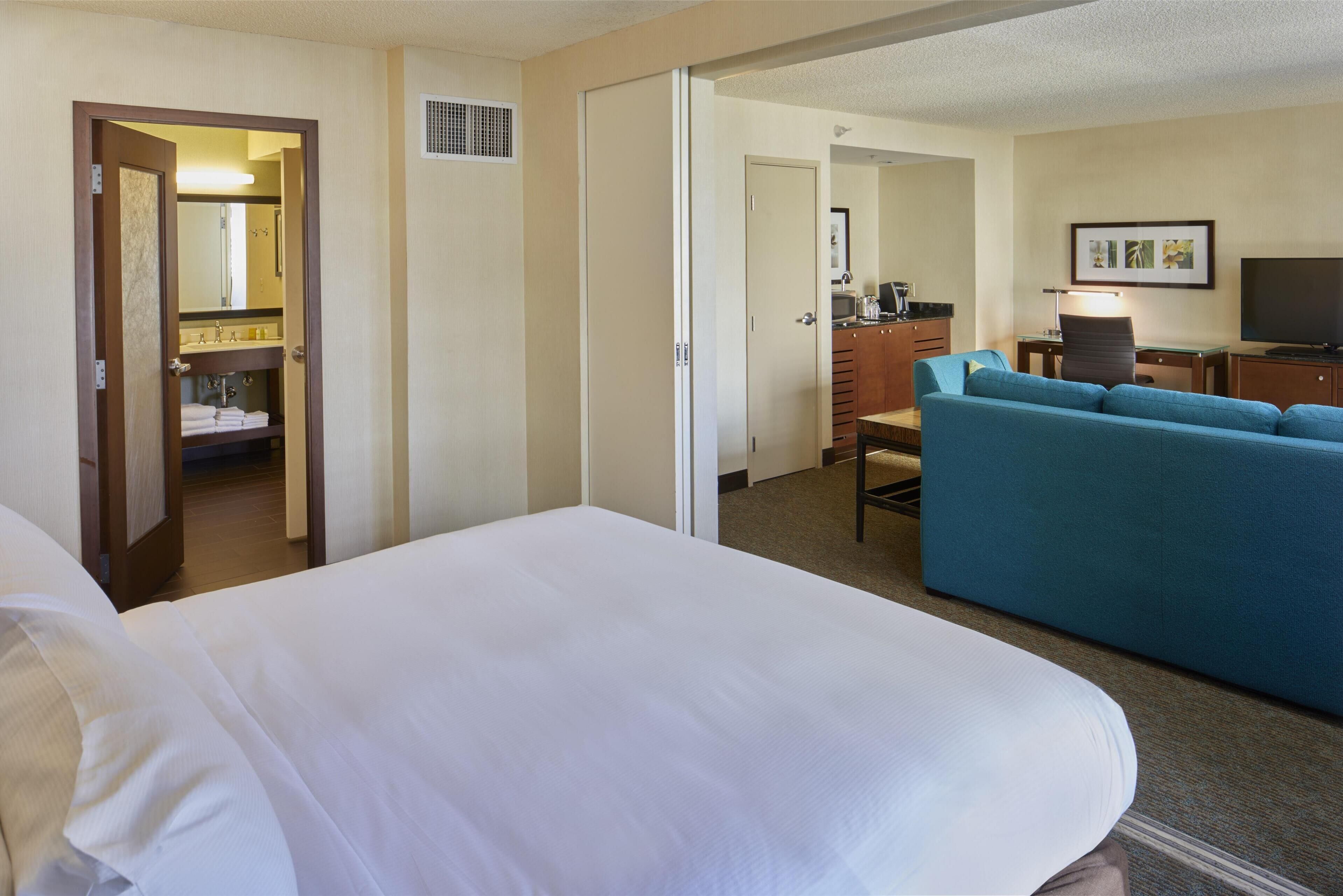 Quarto de hotel com cama, sofá azul, escrivaninha com cadeira, televisão e banheiro visível através de uma porta aberta.