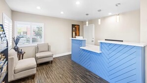 Un área de recepción con mostrador azul, dos sillones beige, una mesita con un florero y una ventana con persianas.
