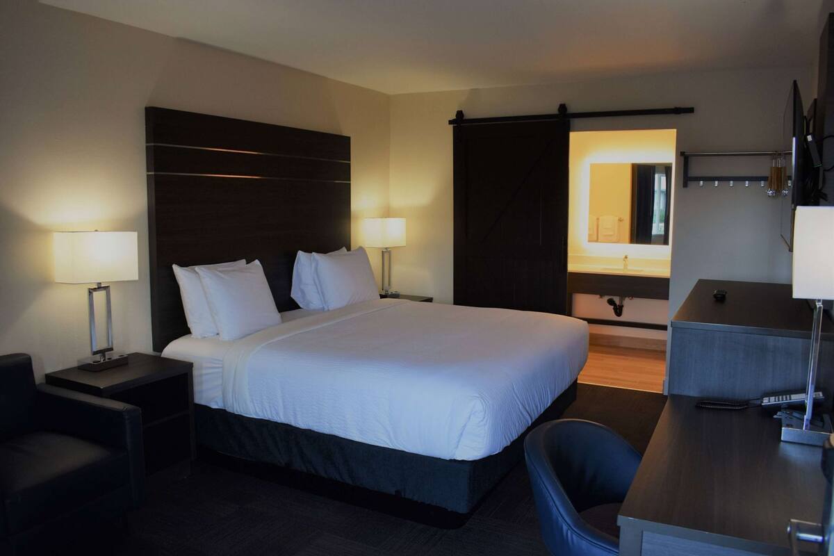 A hotel room with a large bed, bedside tables with lamps, a desk with a chair, a wardrobe, and a bathroom with a mirror.