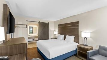 A hotel room with a large bed, a desk with a computer, a TV, a sliding door, and a nightstand with a lamp.