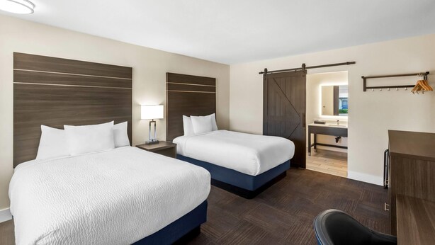 Habitación de hotel con dos camas, un escritorio, una silla, un espejo y un baño visible a través de una puerta abierta.