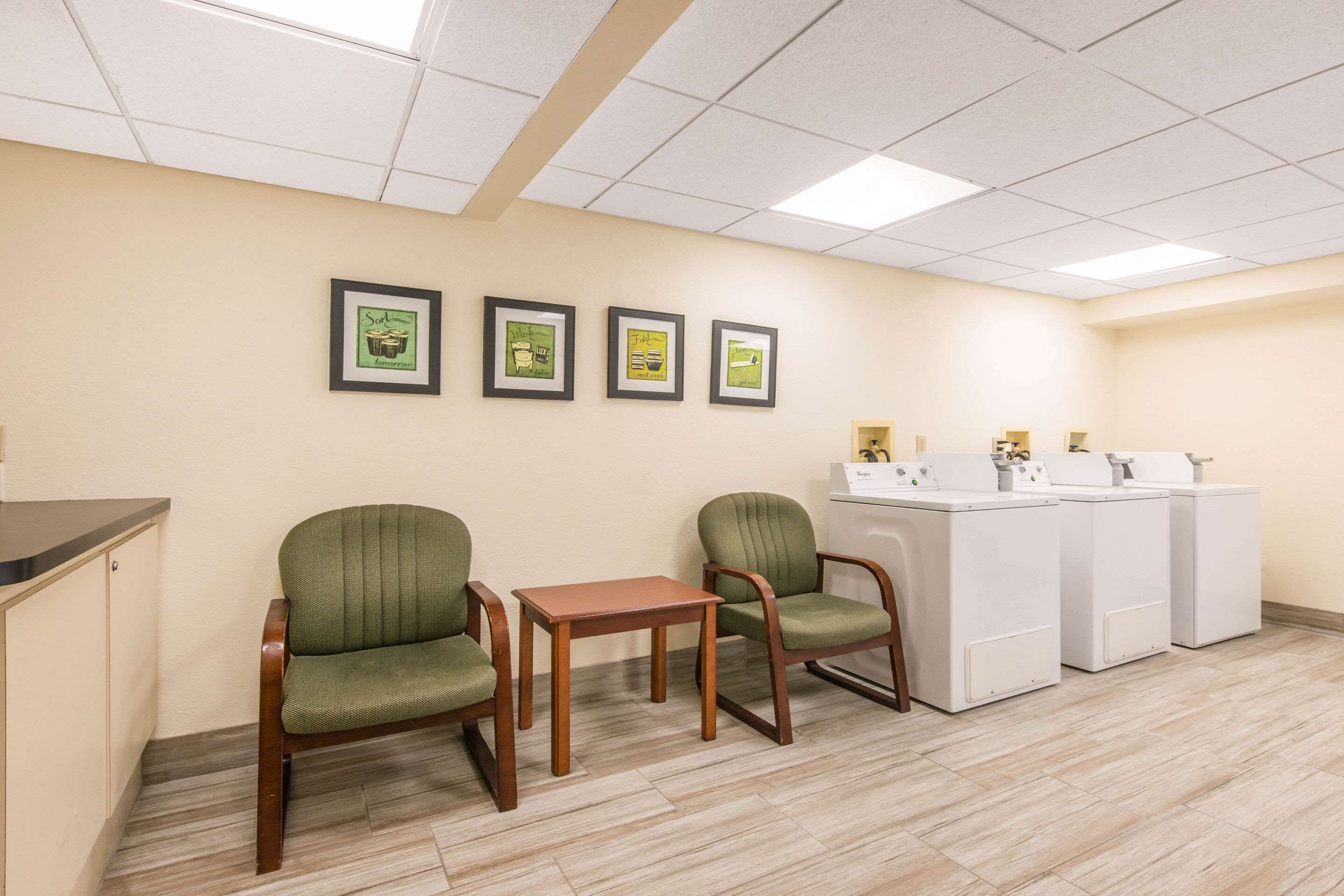 A room with two green chairs, a wooden table, and a row of white washing machines.