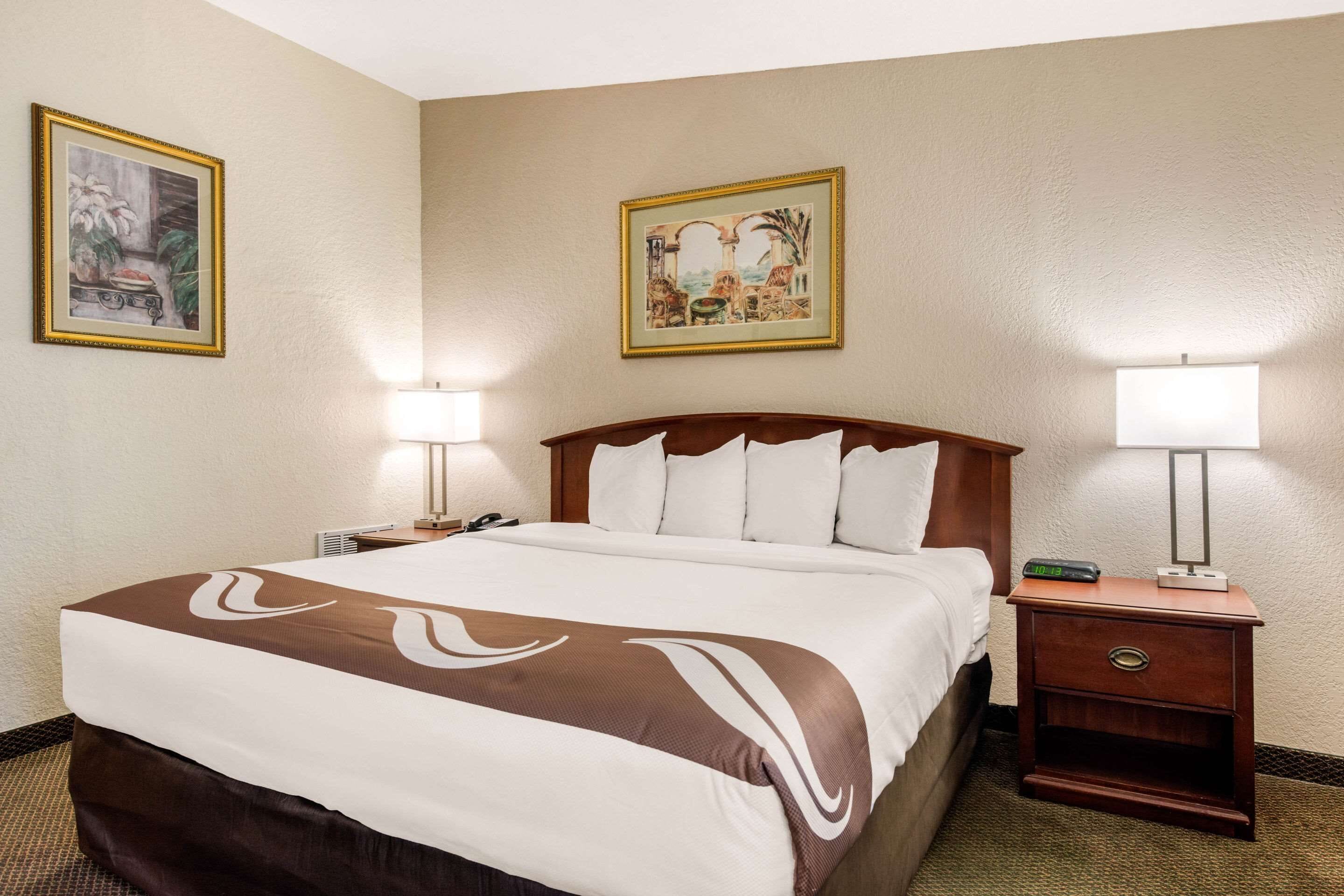 Une chambre d’hôtel avec un grand lit, deux tables de chevet, une petite table, une lampe et deux tableaux encadrés accrochés aux murs.