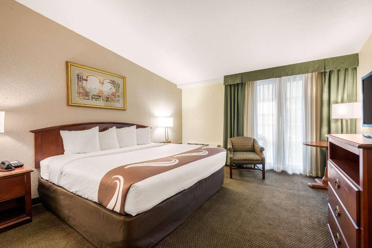 Una habitación de hotel con una cama grande, una silla, un escritorio con lámpara, un cuadro en la pared y un televisor sobre un aparador.