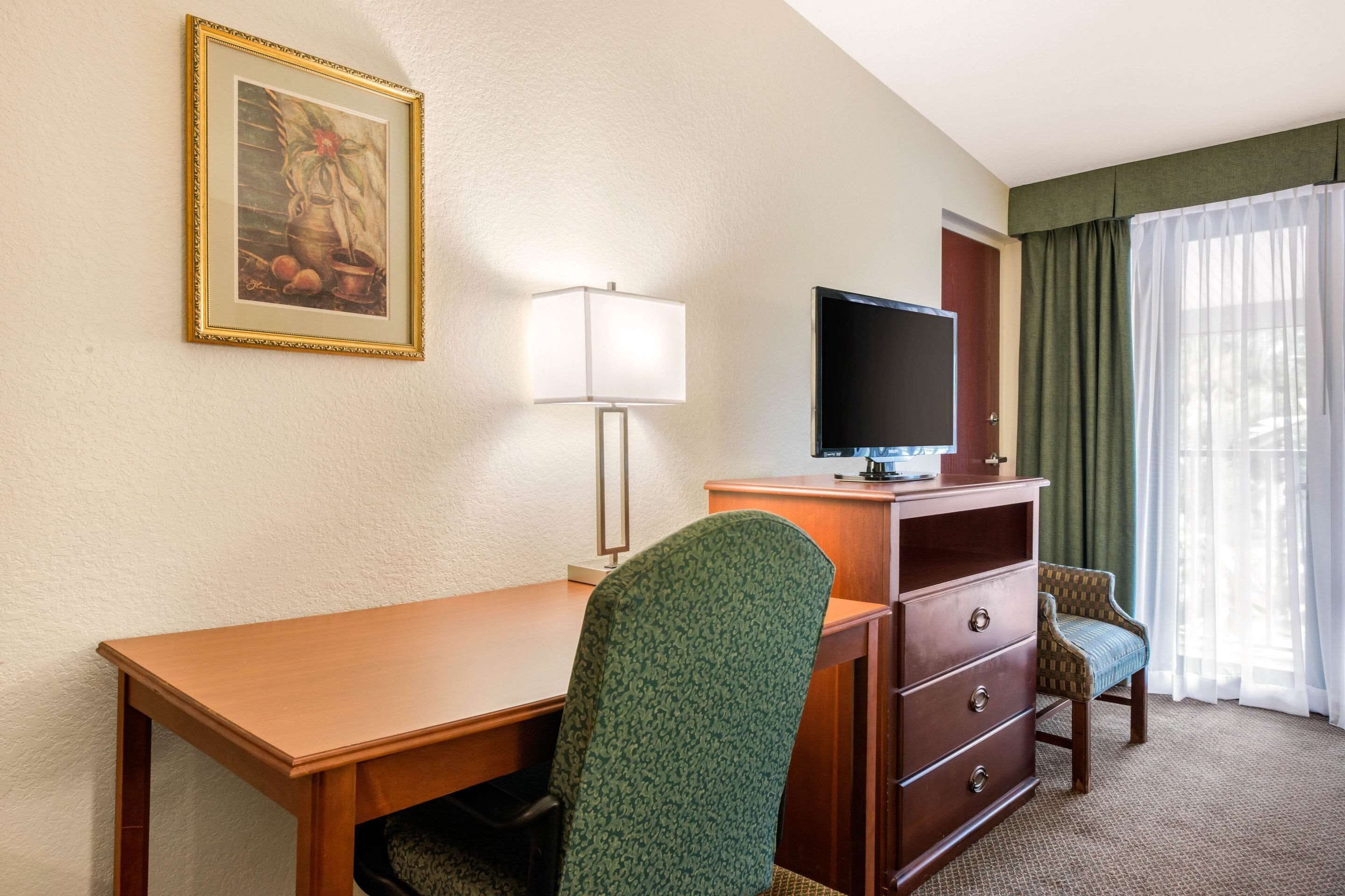 Quarto de hotel com uma escrivaninha de madeira, uma poltrona estofada em verde, uma televisão sobre um móvel de madeira, um quadro emoldurado na parede e uma janela com cortinas leves.
