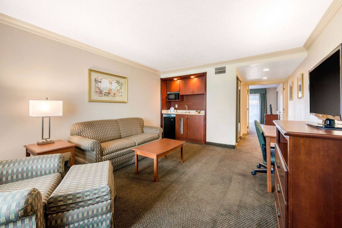 Habitación de hotel con sala de estar que incluye un sofá, un sillón y una mesa de centro. Cuenta con una kitchenette con microondas y armarios, además de un televisor sobre un mueble.