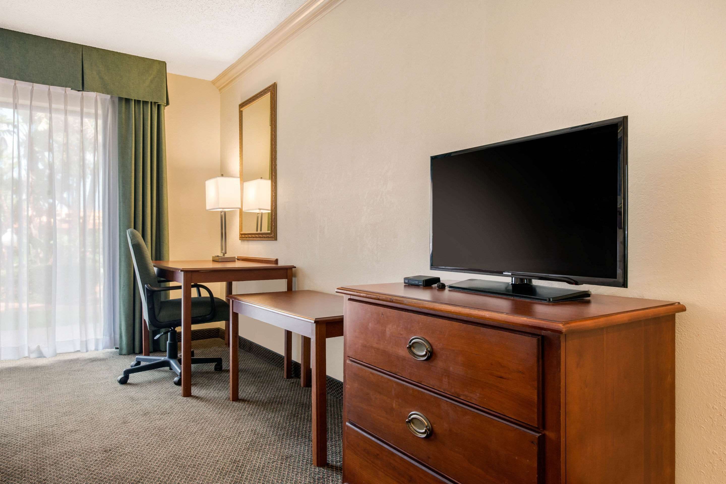 A hotel room with a wooden desk, a chair, a television on a dresser, a lamp, and a large window with curtains.