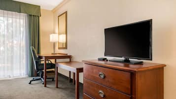 A hotel room with a wooden desk, a chair, a television on a dresser, a lamp, and a large window with curtains.