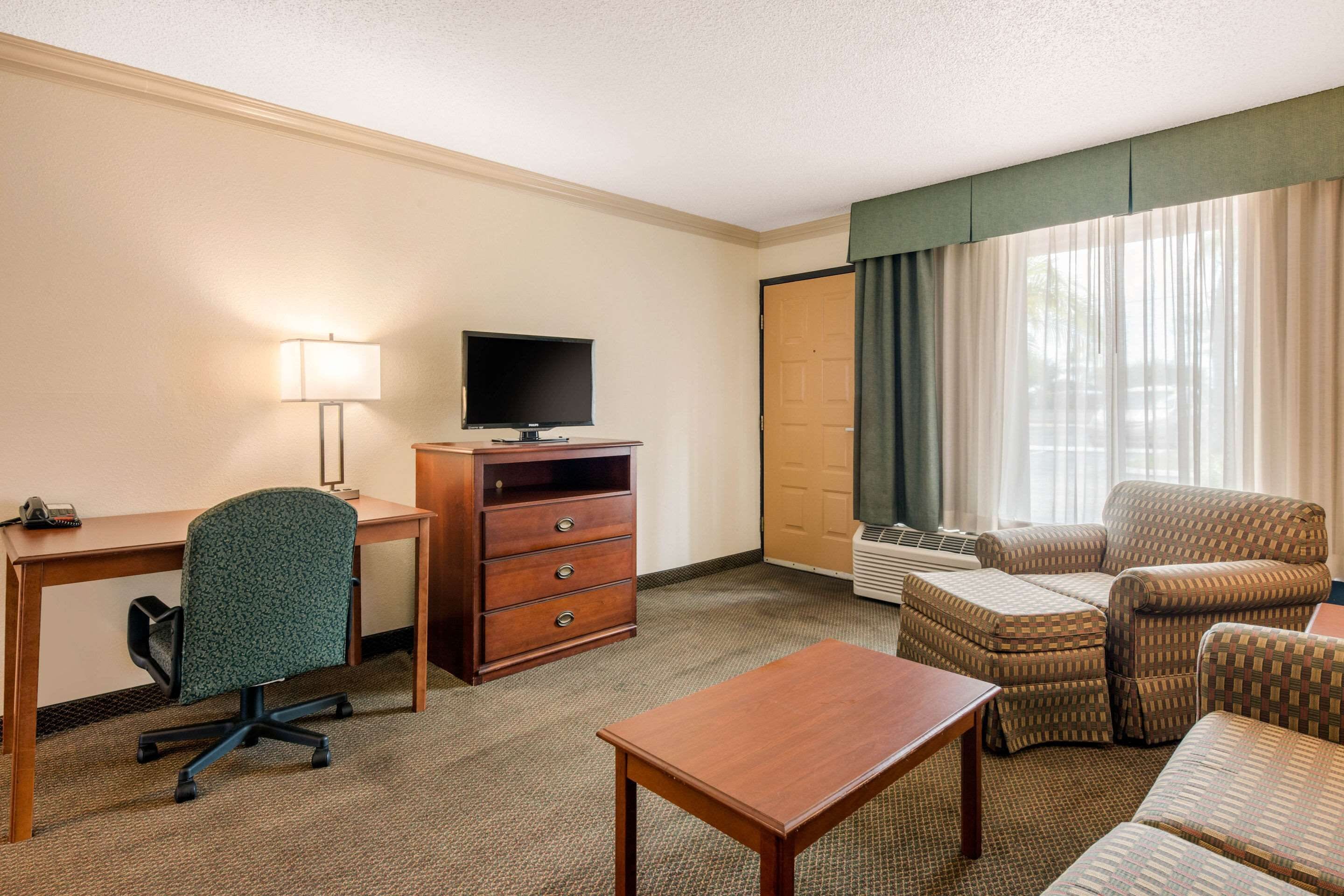 A hotel room with a desk, chair, television, and a seating area with a sofa and ottoman.