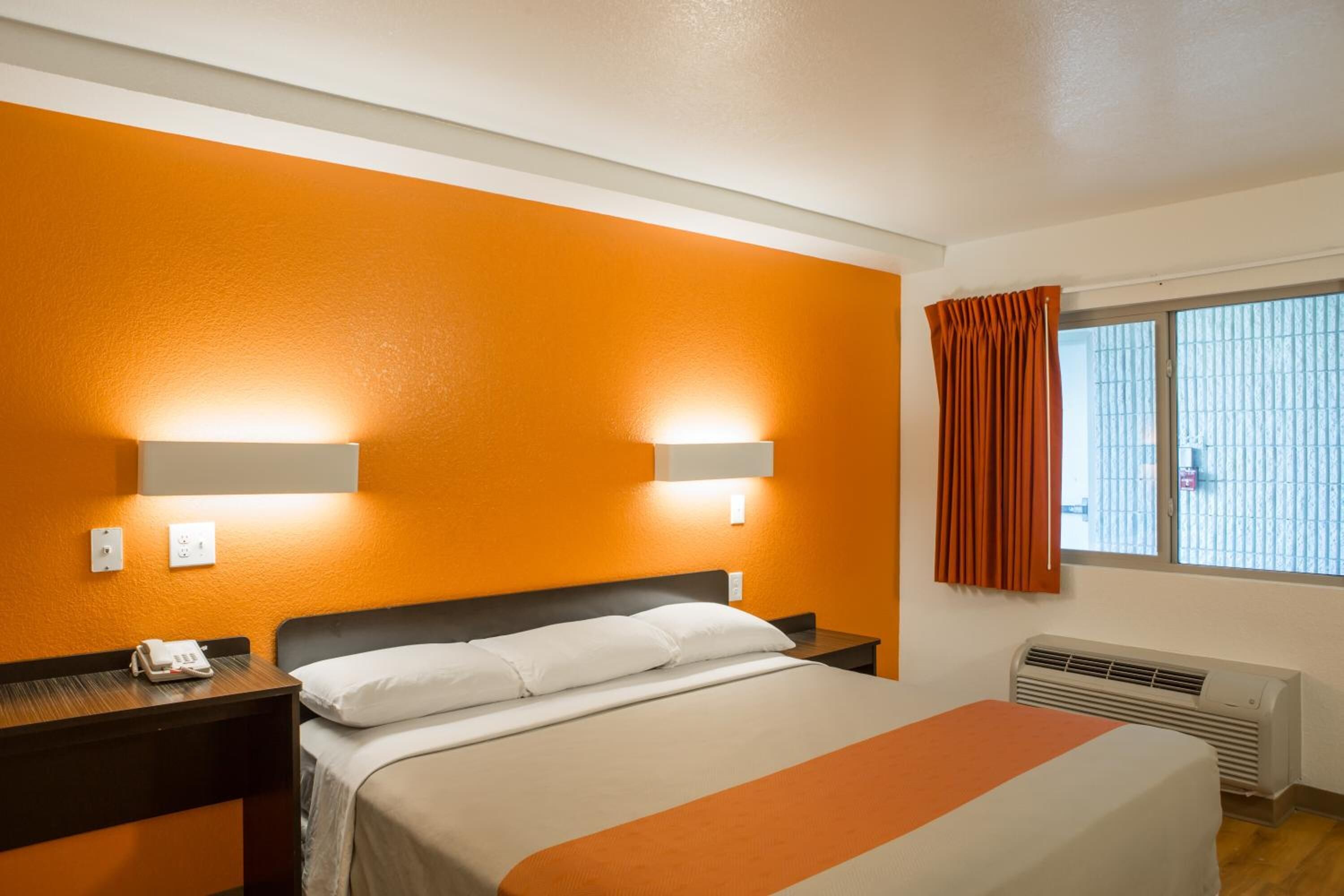 A hotel room with an orange accent wall, a bed with white bedding, a nightstand with a phone, and a window with curtains.
