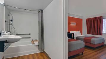 A modern bathroom with a sink, shower, and toilet. Adjacent is a bedroom with two beds and a window.