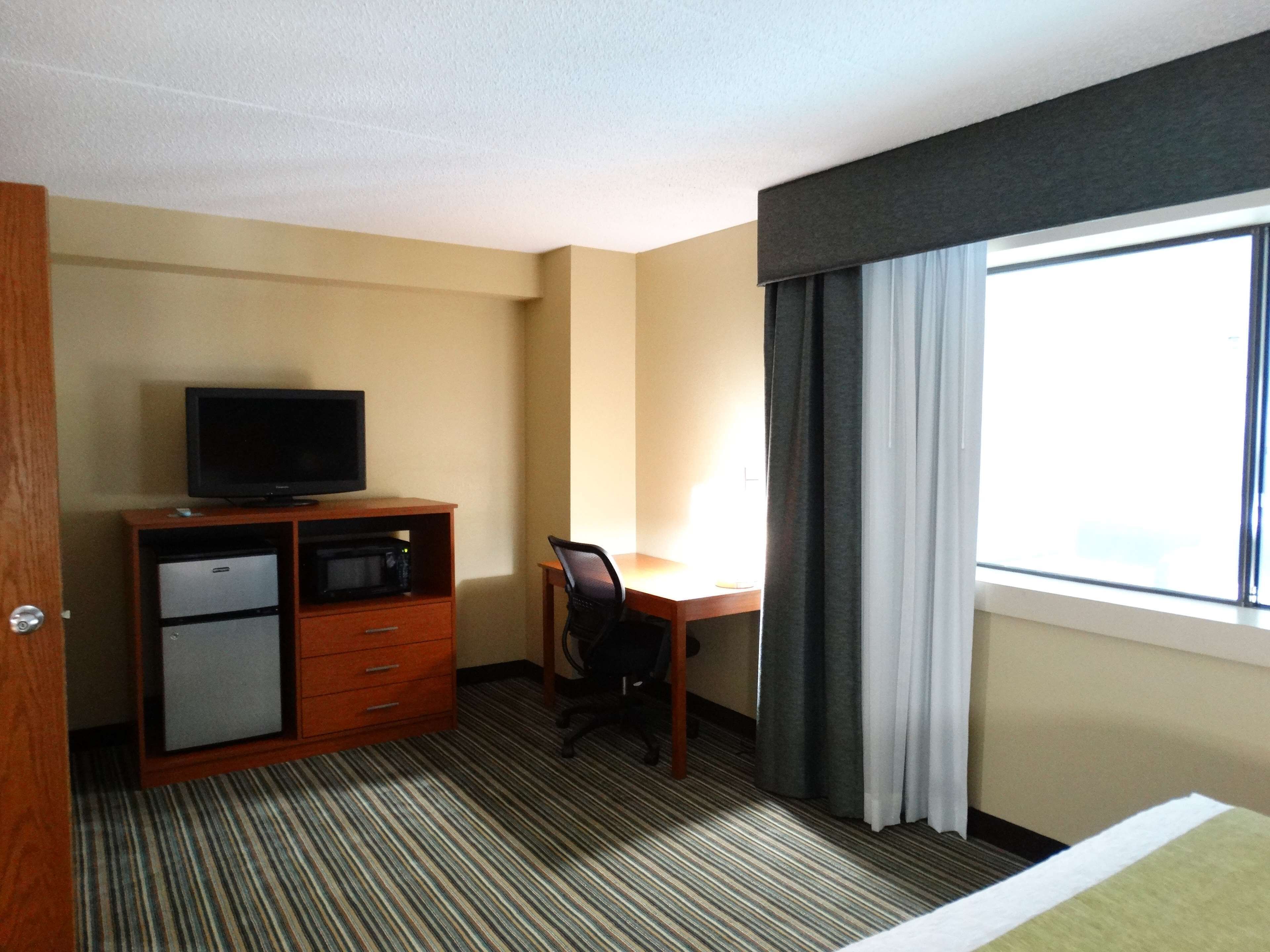 A hotel room with a bed, a desk with a chair, a television on a cabinet, a small refrigerator, and a window with curtains.