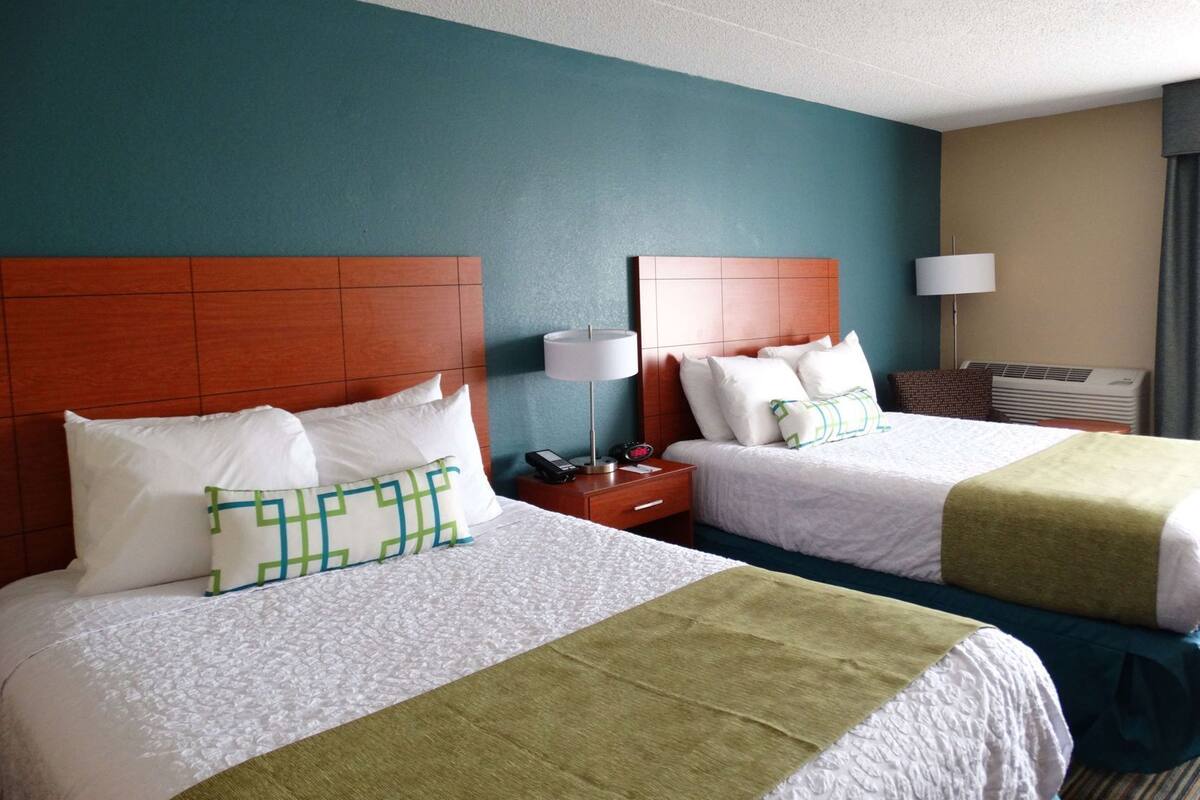 A hotel room with two beds, a nightstand with a lamp, a telephone, and an air conditioning unit.