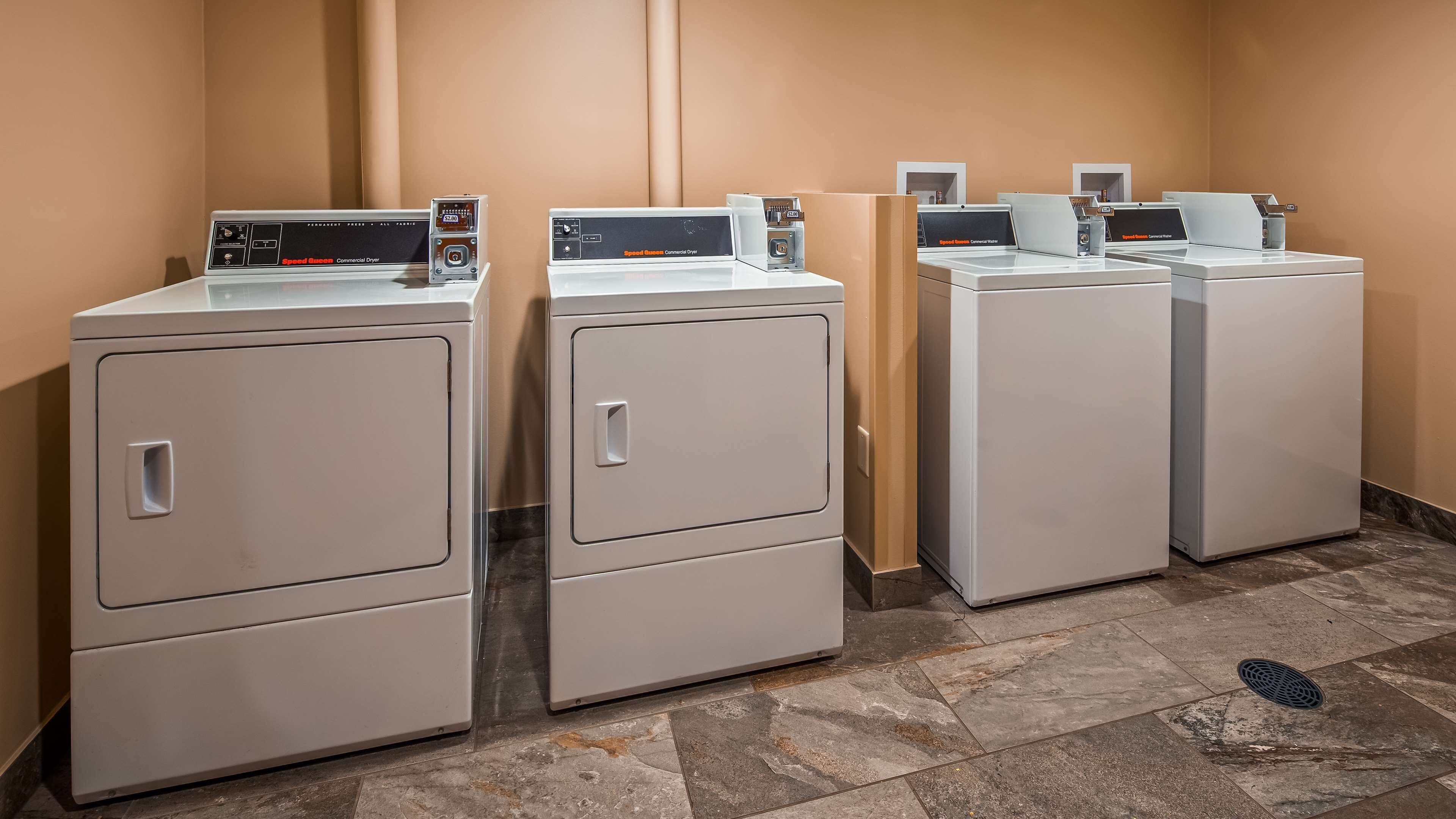 A row of commercial dryers in a public area, likely a laundromat, with a tiled floor and beige walls.