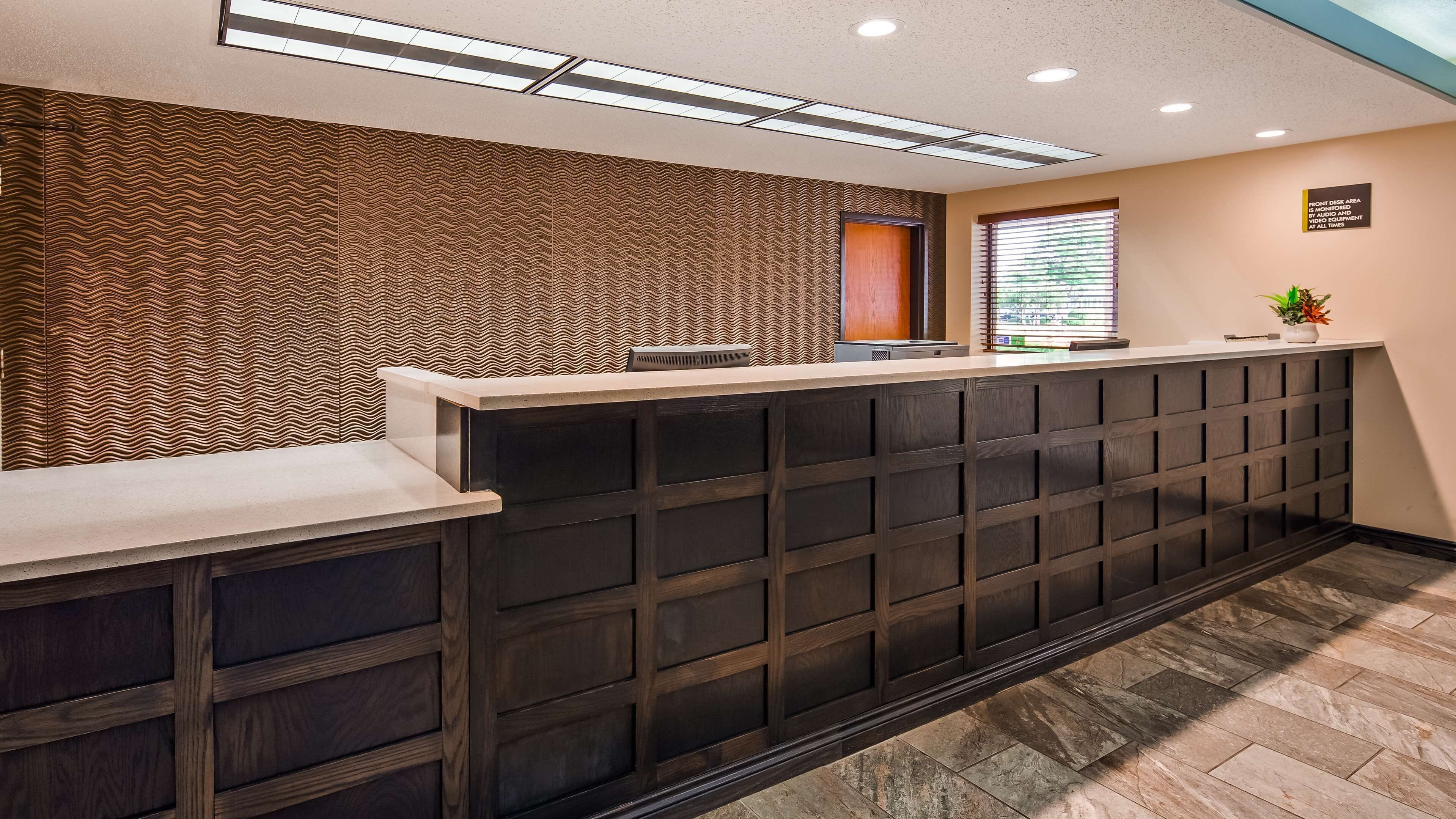 Un comptoir d'accueil avec un mur décoré, une fenêtre avec des stores et un panneau indiquant l'emplacement du bureau d'accueil.