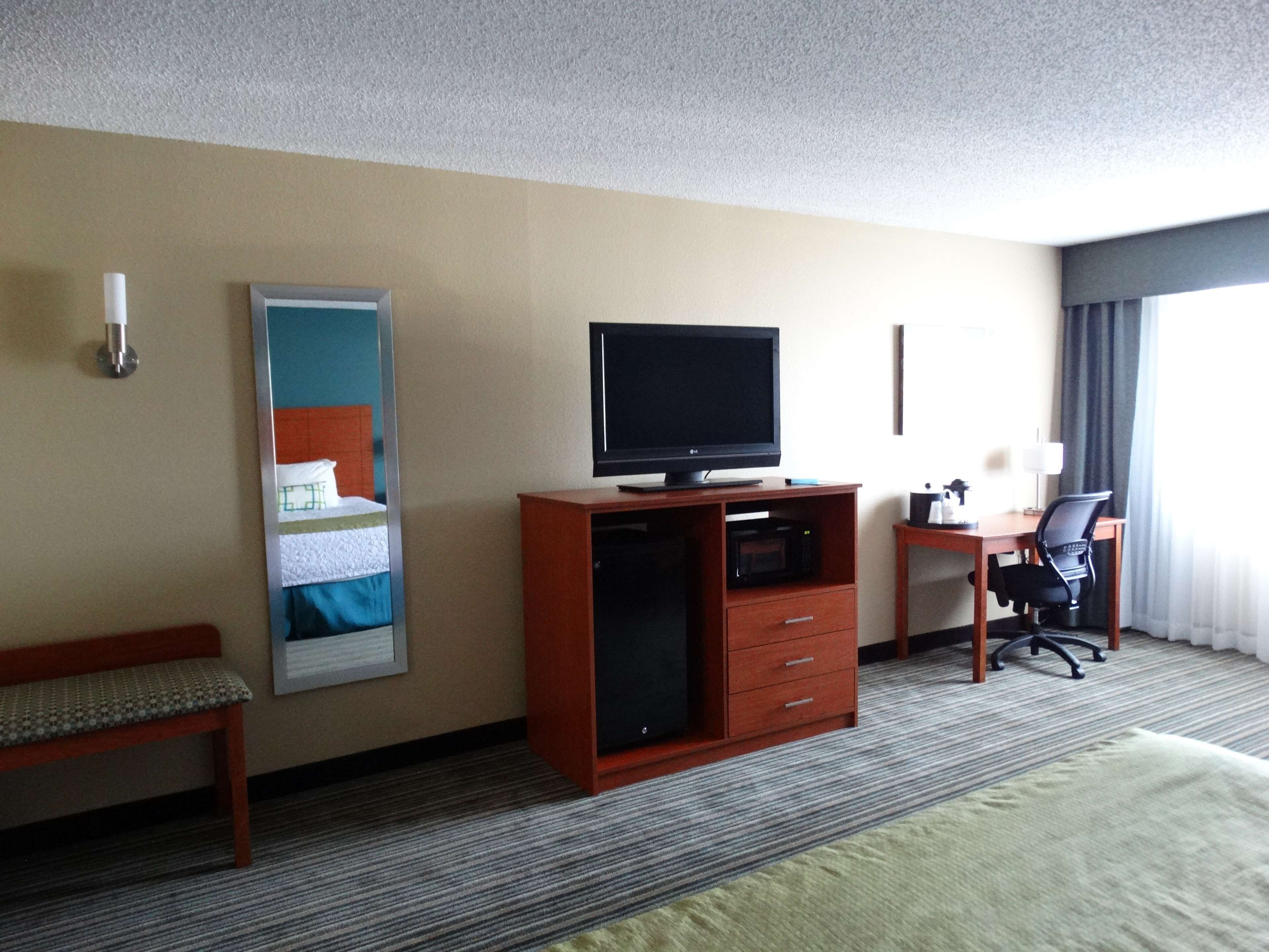 A hotel room with a bed, a desk with a chair, a television on a wooden cabinet, a bedside table with a lamp, and a window with curtains.