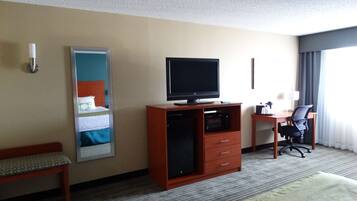 A hotel room with a bed, a desk with a chair, a television on a wooden cabinet, a bedside table with a lamp, and a window with curtains.