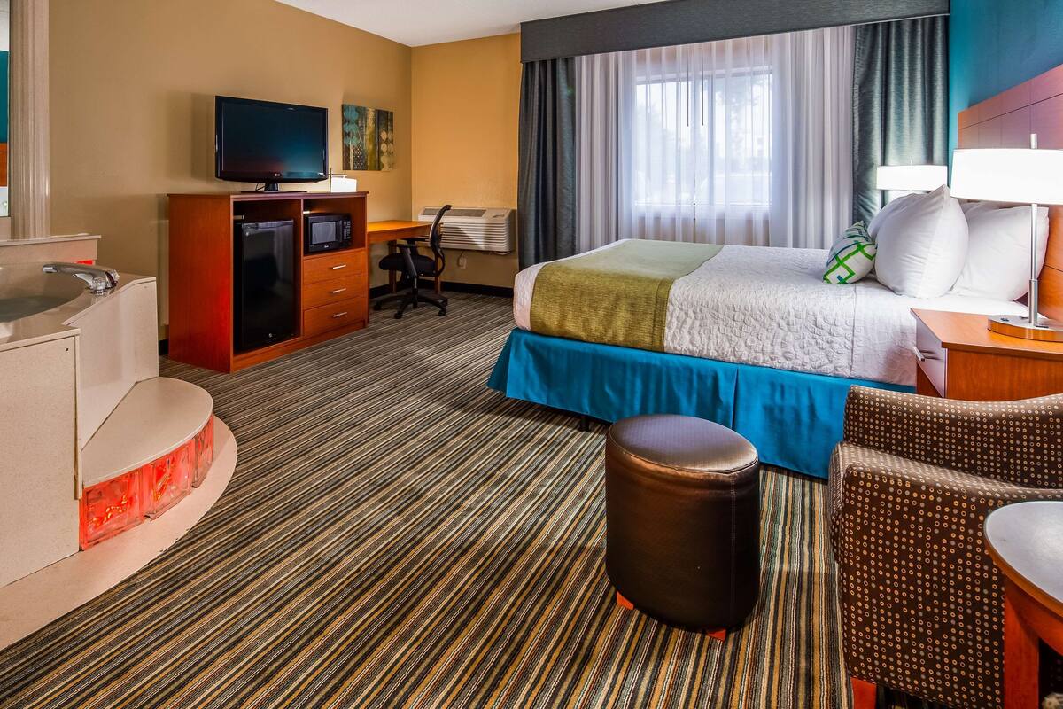 A hotel room with a bed, a desk, a chair, a TV, a minibar, and a bathroom with a bathtub.