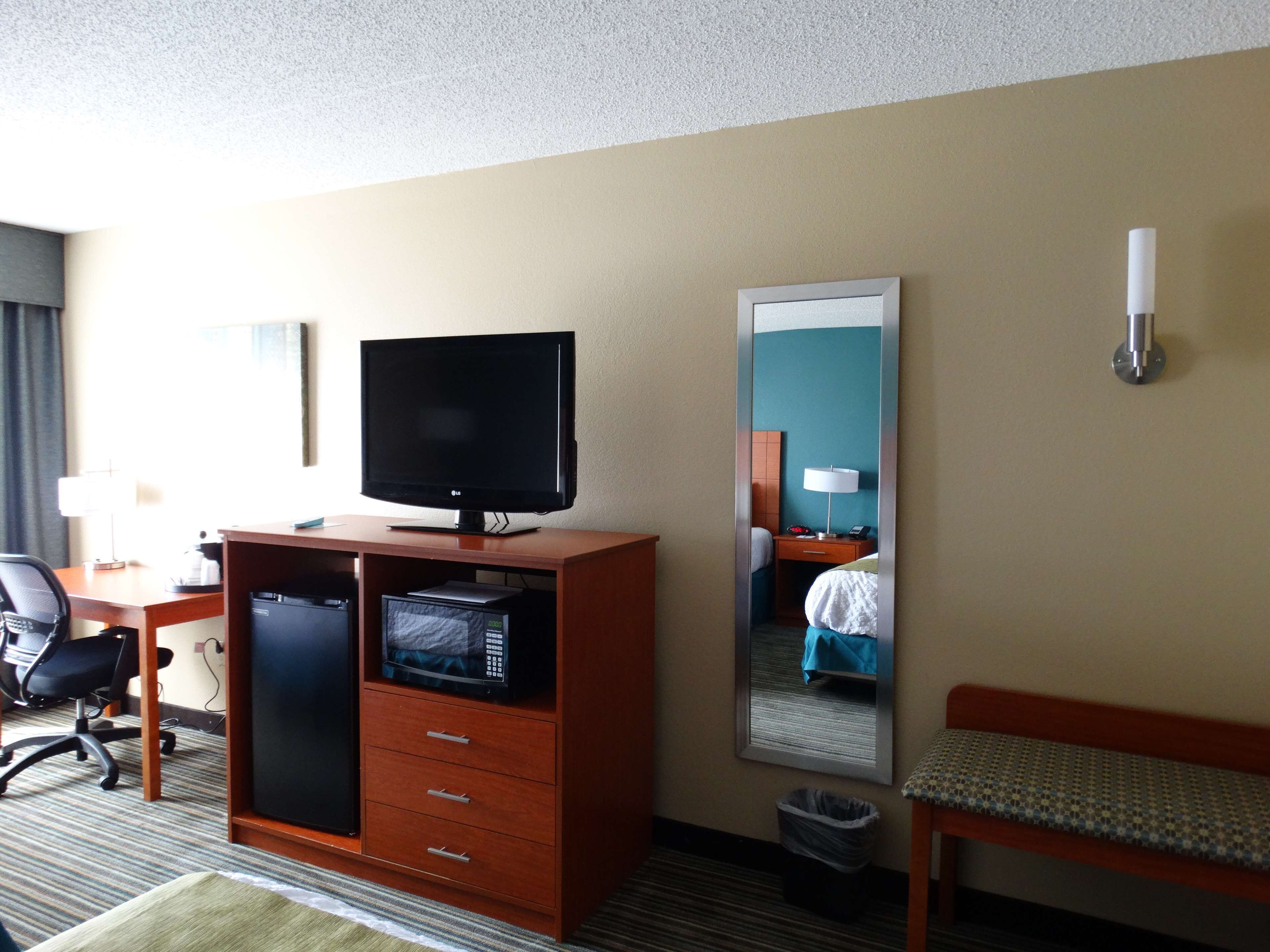A hotel room with a television on a wooden cabinet, a desk with a chair, a bed with a blue blanket, a mirror, and a trash bin.
