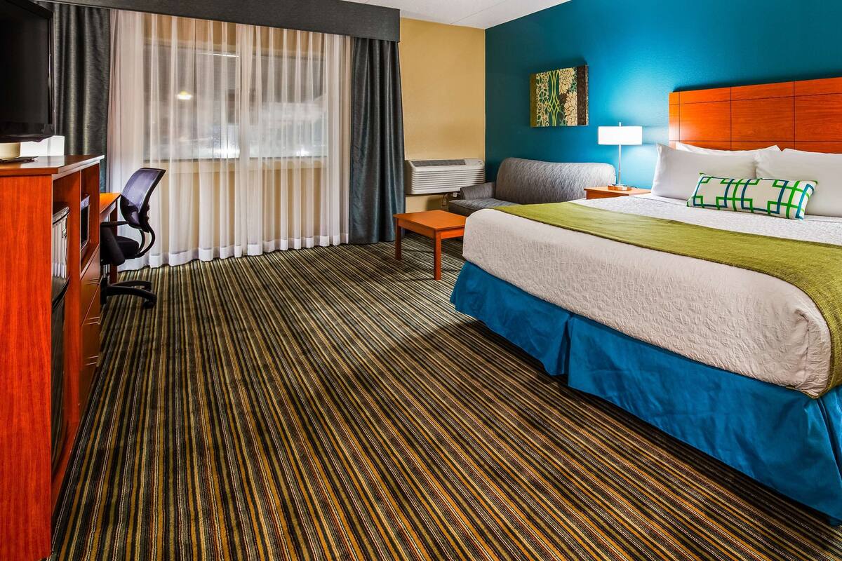 A hotel room with a bed, a desk, a chair, a window with curtains, and a wall with a painting.
