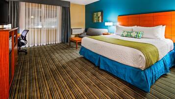 A hotel room with a bed, a desk, a chair, a window with curtains, and a wall with a painting.