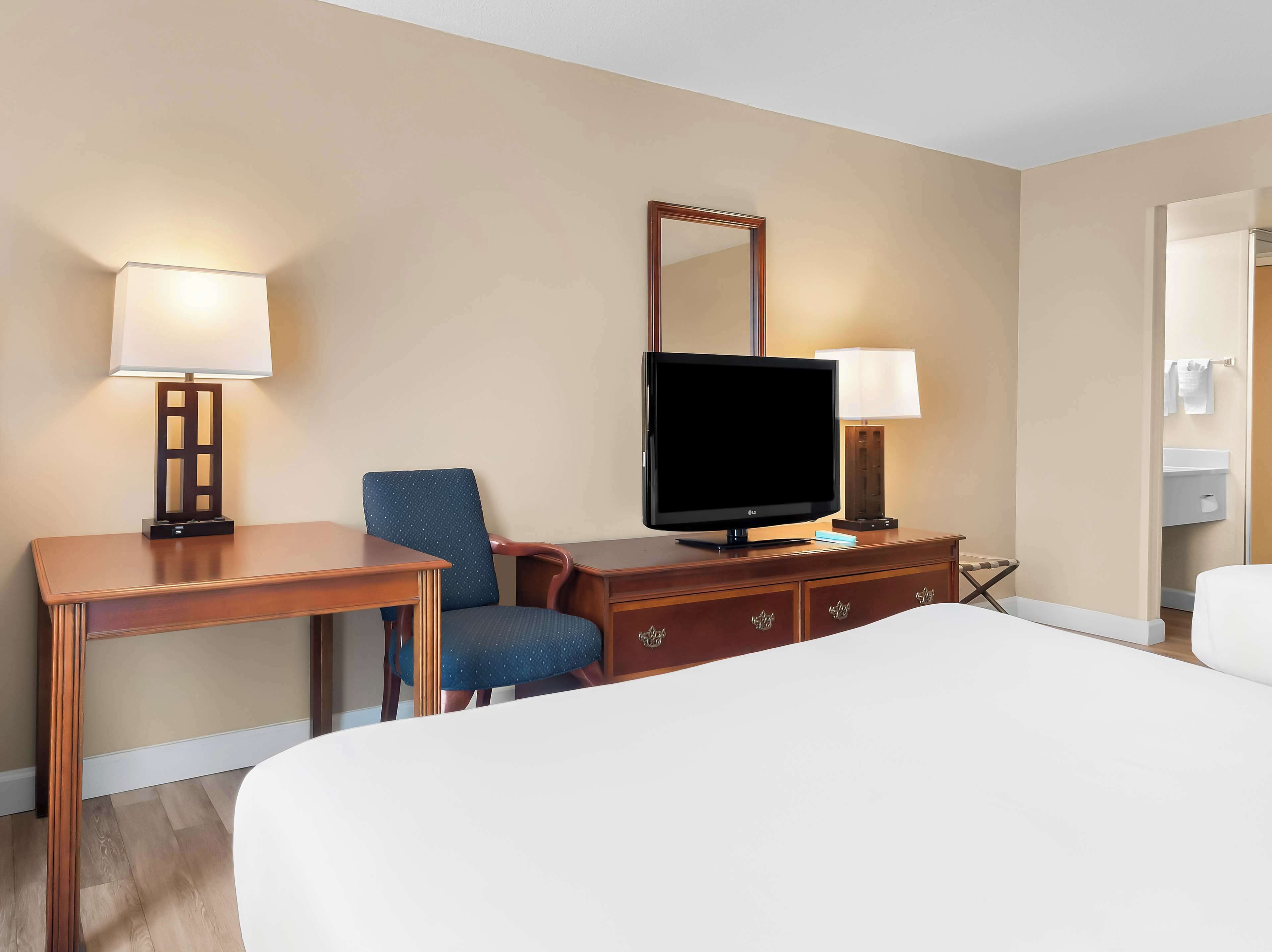 A hotel room with a bed, a desk with a lamp, a television, a chair, and a dresser.