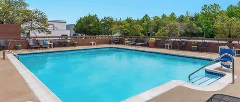 A rectangular swimming pool with a tiled edge, surrounded by lounge chairs and tables, and a brick wall in the background.