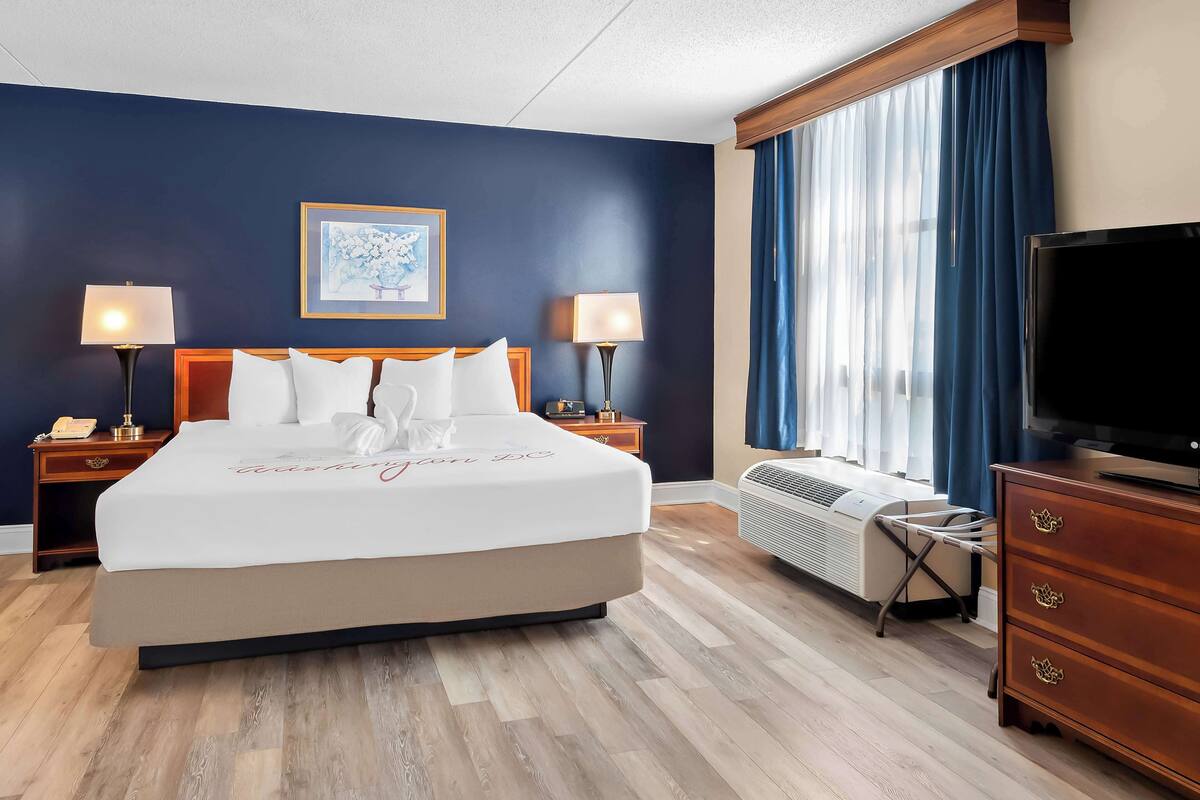 A hotel room with a large bed, two bedside lamps, a flat-screen TV, a dresser, and a window with curtains.