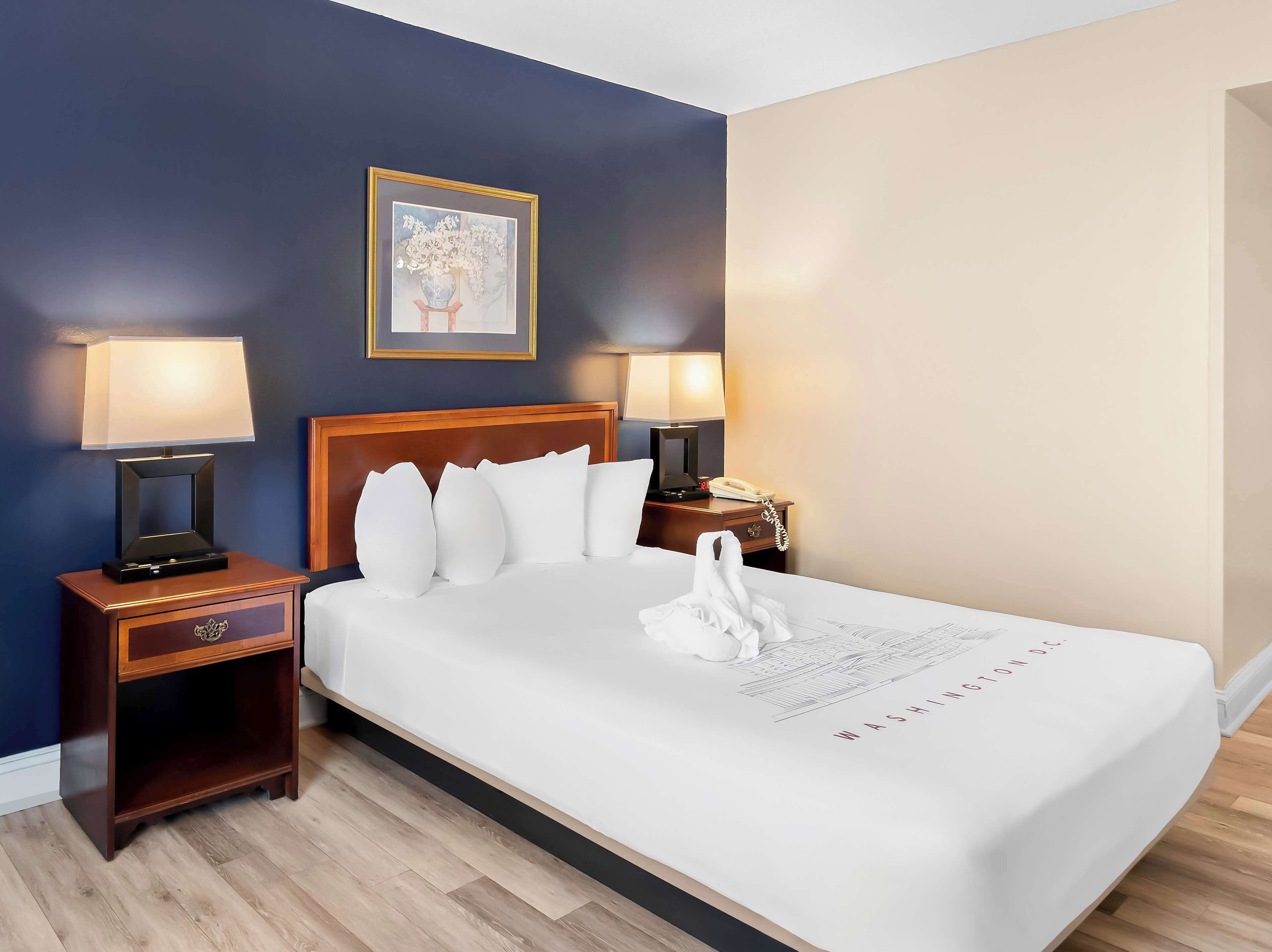 A hotel room with a bed, two bedside lamps, a nightstand, a framed picture on the wall, and a white swan decoration on the bed.