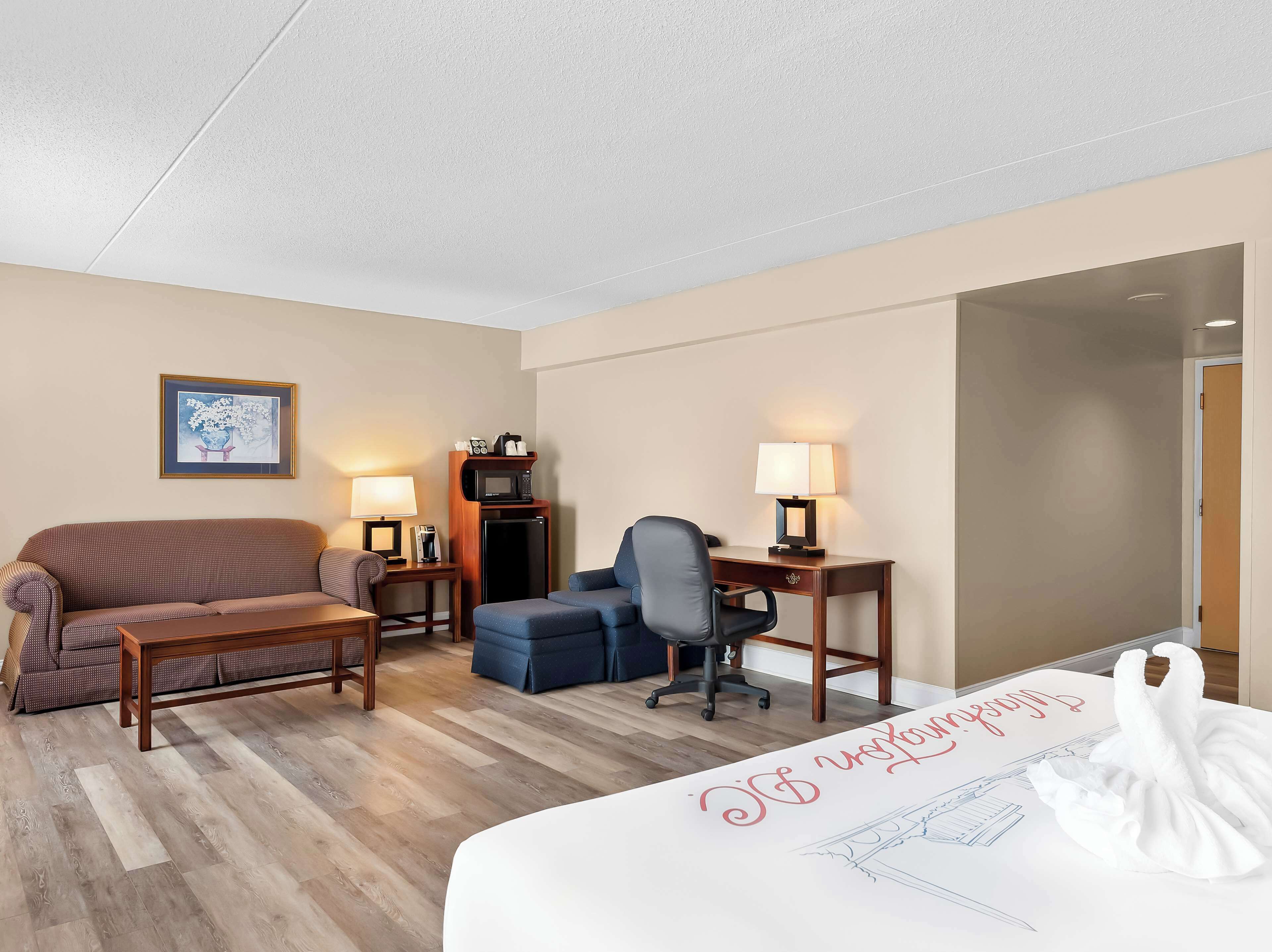 A hotel room with a bed, a desk, a chair, a sofa, a microwave, a coffee maker, and a framed picture on the wall.