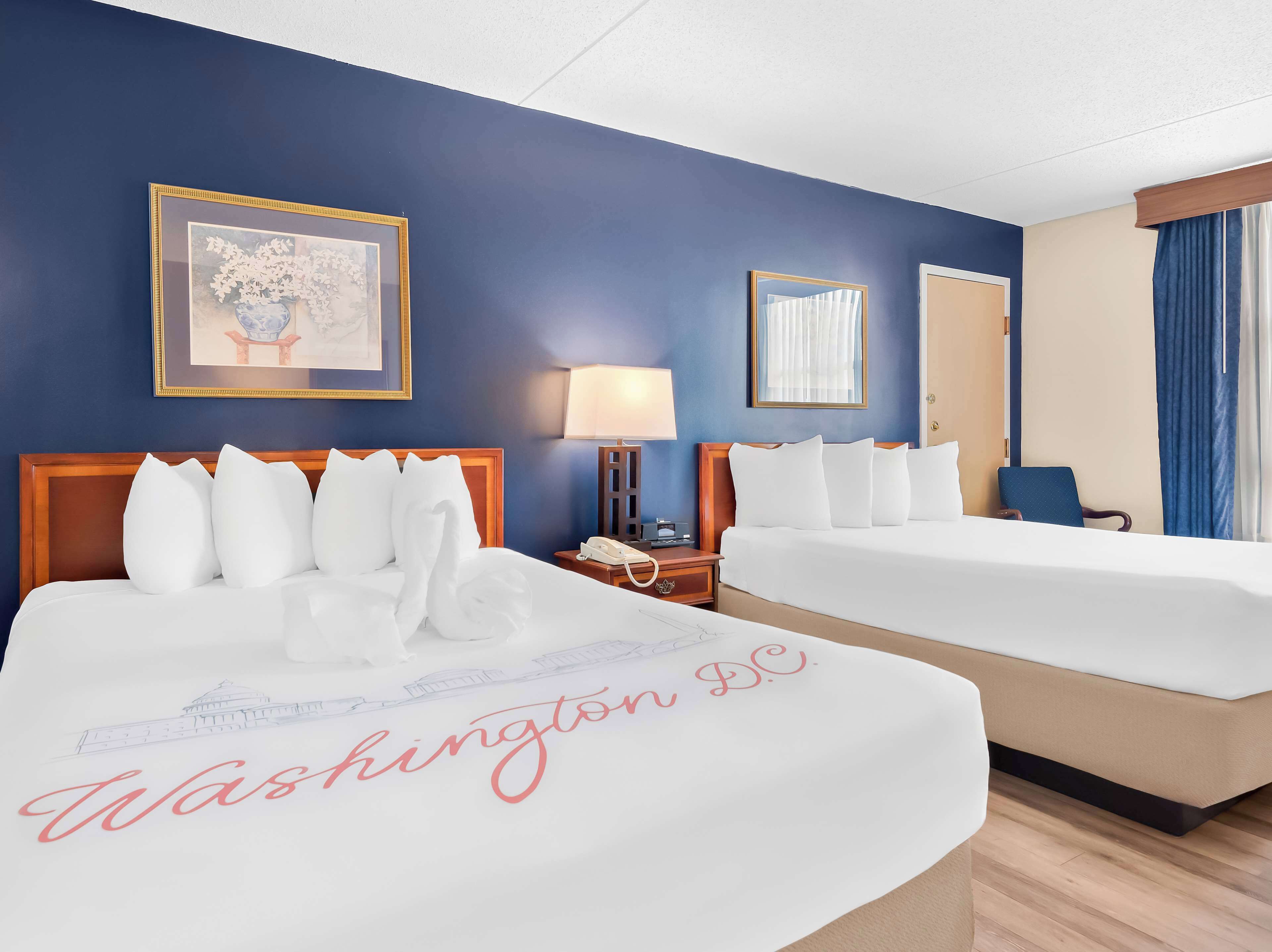 Habitación de hotel con dos camas, una mesita de noche con lámpara, una silla y cuadros en las paredes.