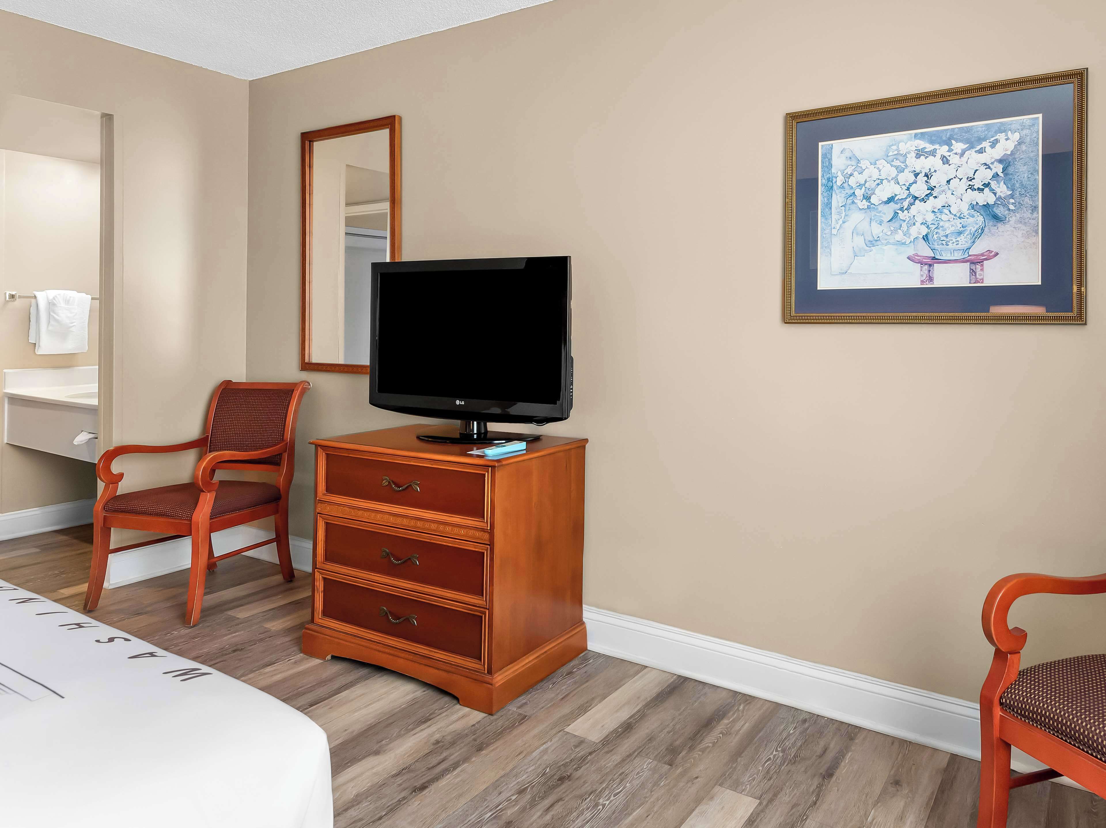 A hotel room with a wooden dresser, a television, a chair, a mirror, and a framed picture on the wall.